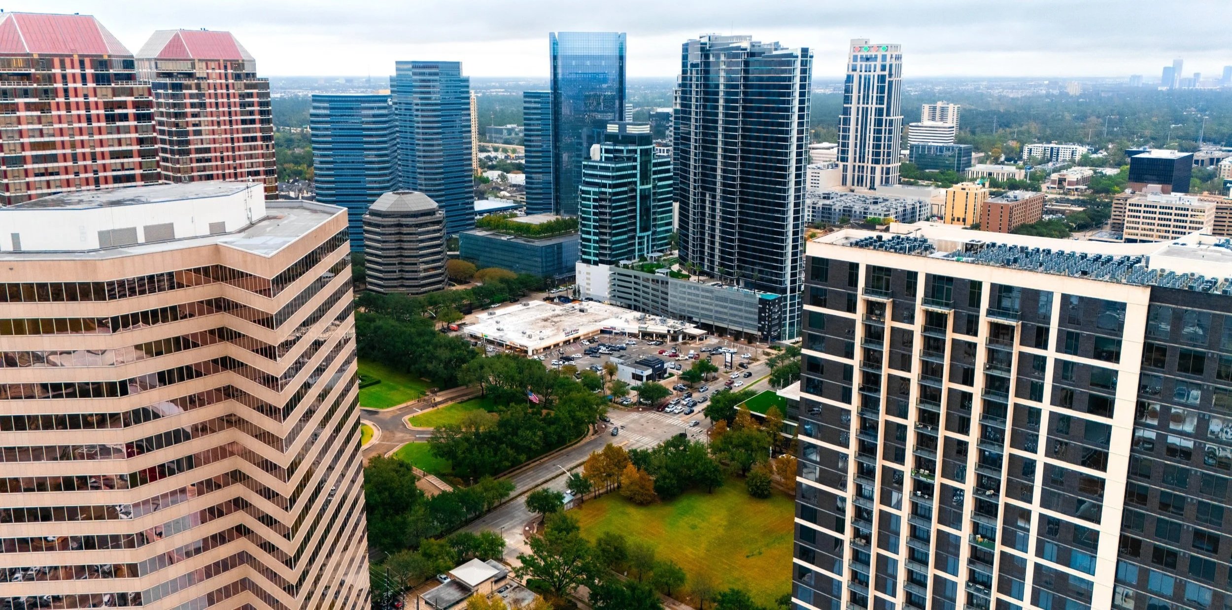 Aerial view of a city skyline with tall modern buildings, green trees, and a parking lot.