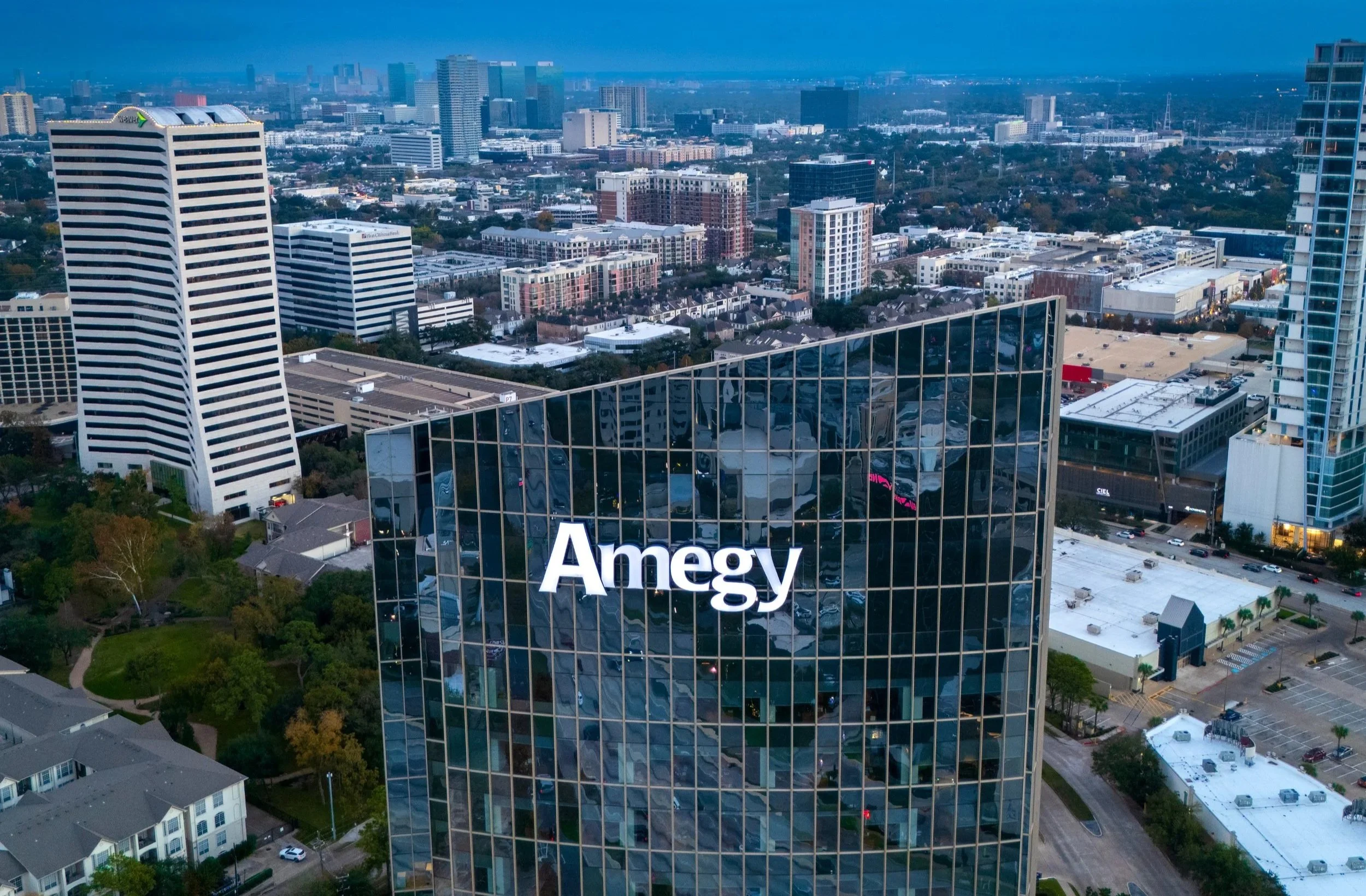 Aerial view of a cityscape with modern buildings, including a prominent glass office building with the 'Ameg' logo, reflecting surrounding structures, during dusk.