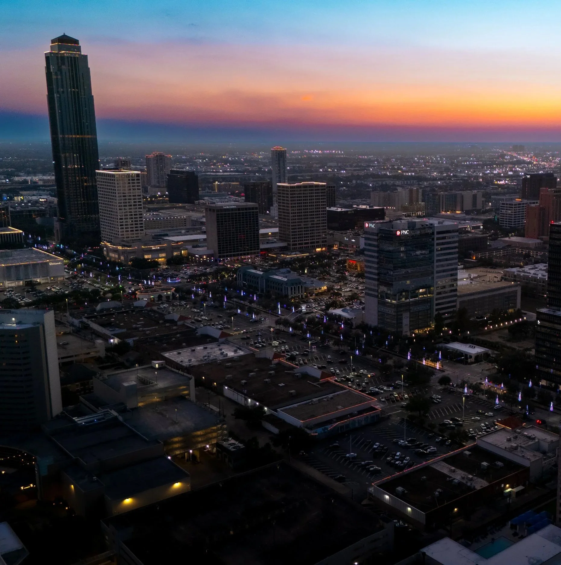 City skyline at sunset with high-rise buildings, parking lots, streets, and colorful sky in the background.