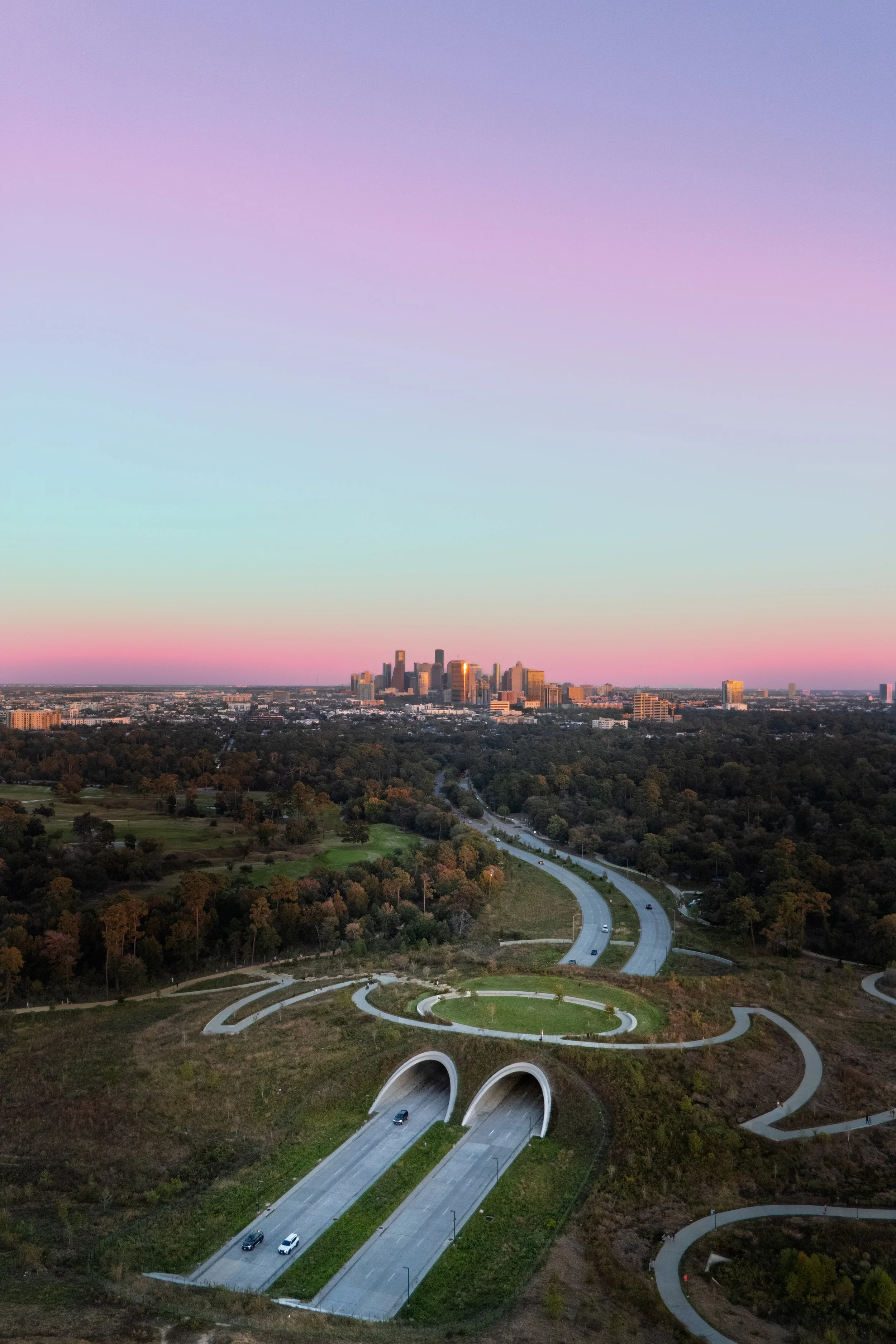 An aerial view of a city skyline during sunset, with a highway leading into tunnels in the foreground, surrounded by trees and greenery.