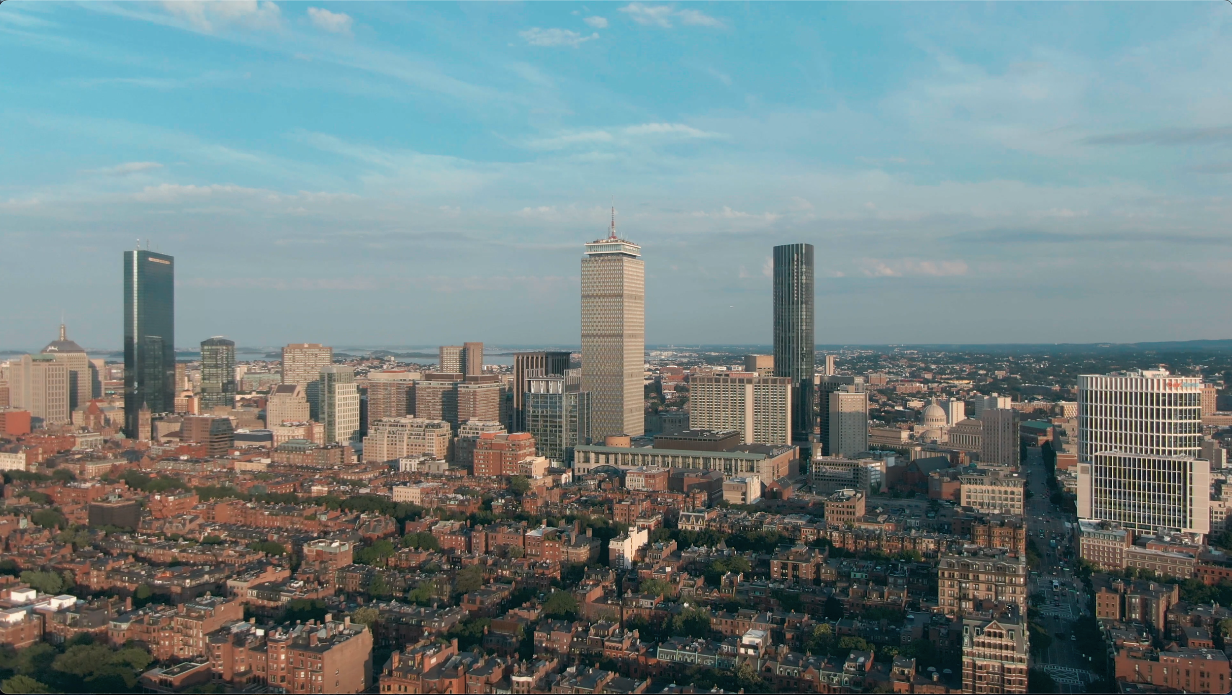 Aerial view of Boston skyline with tall skyscrapers, historic brick buildings, and the Boston Harbor in the distance under a partly cloudy sky.