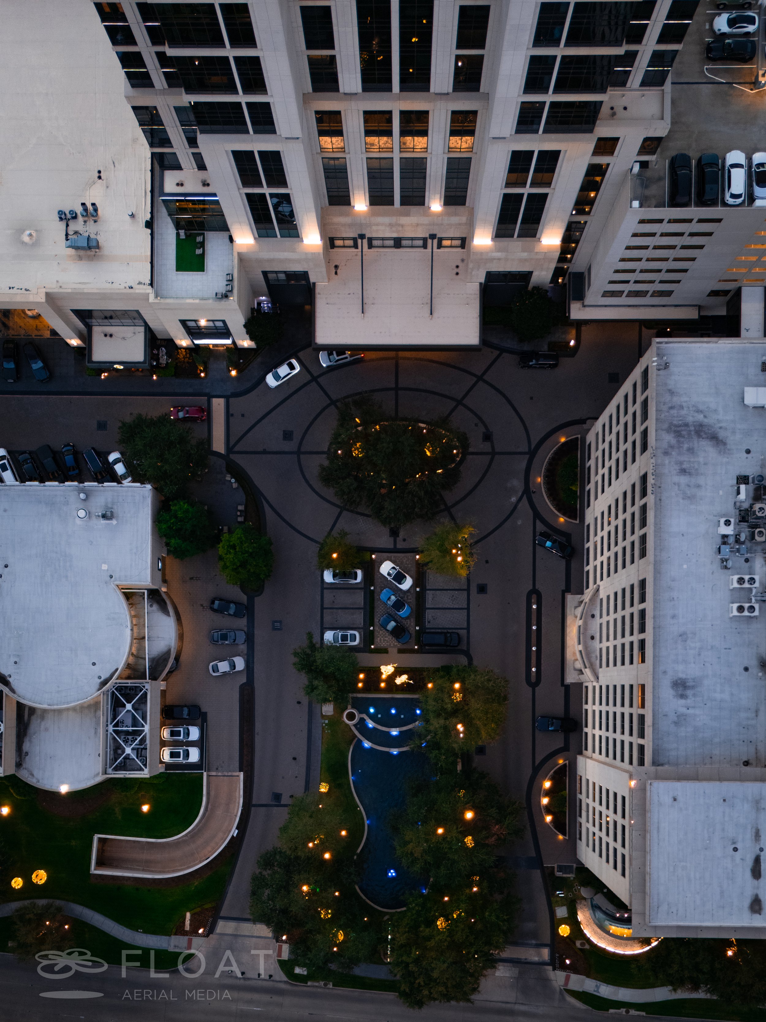 Aerial view of a city courtyard with trees, a fountain, parking spaces, surrounding multi-story buildings, and outdoor lighting at dusk.