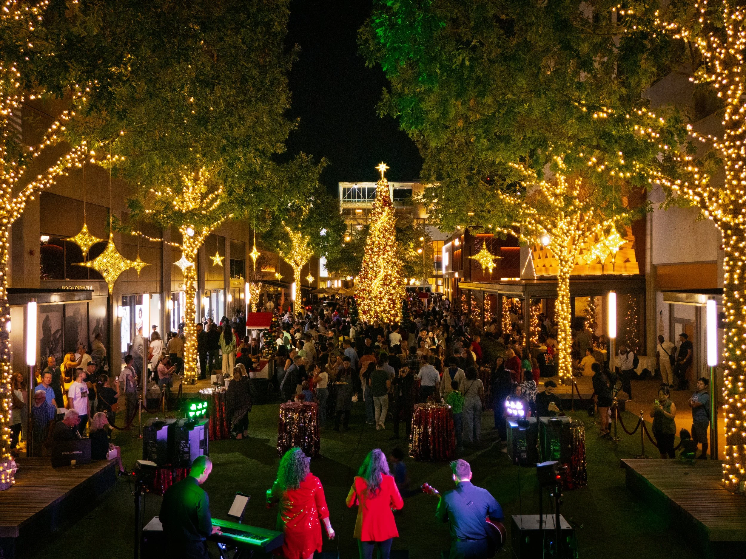 Nighttime outdoor holiday celebration with large decorated Christmas tree, string lights and star ornaments hanging from trees, crowd of people, and live musicians performing on stage.