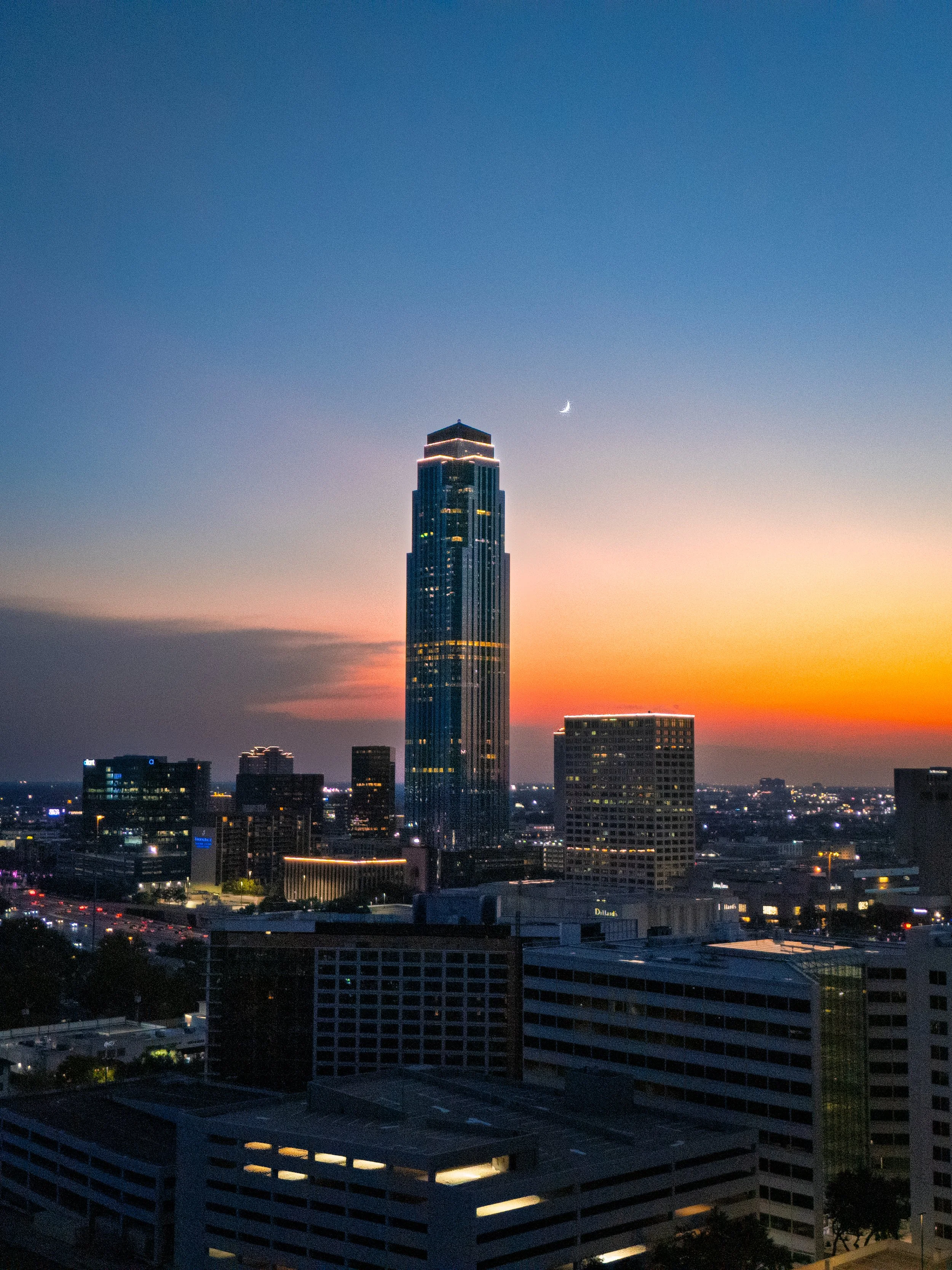 City skyline at sunset with a tall skyscraper, other buildings, and a crescent moon in the sky.