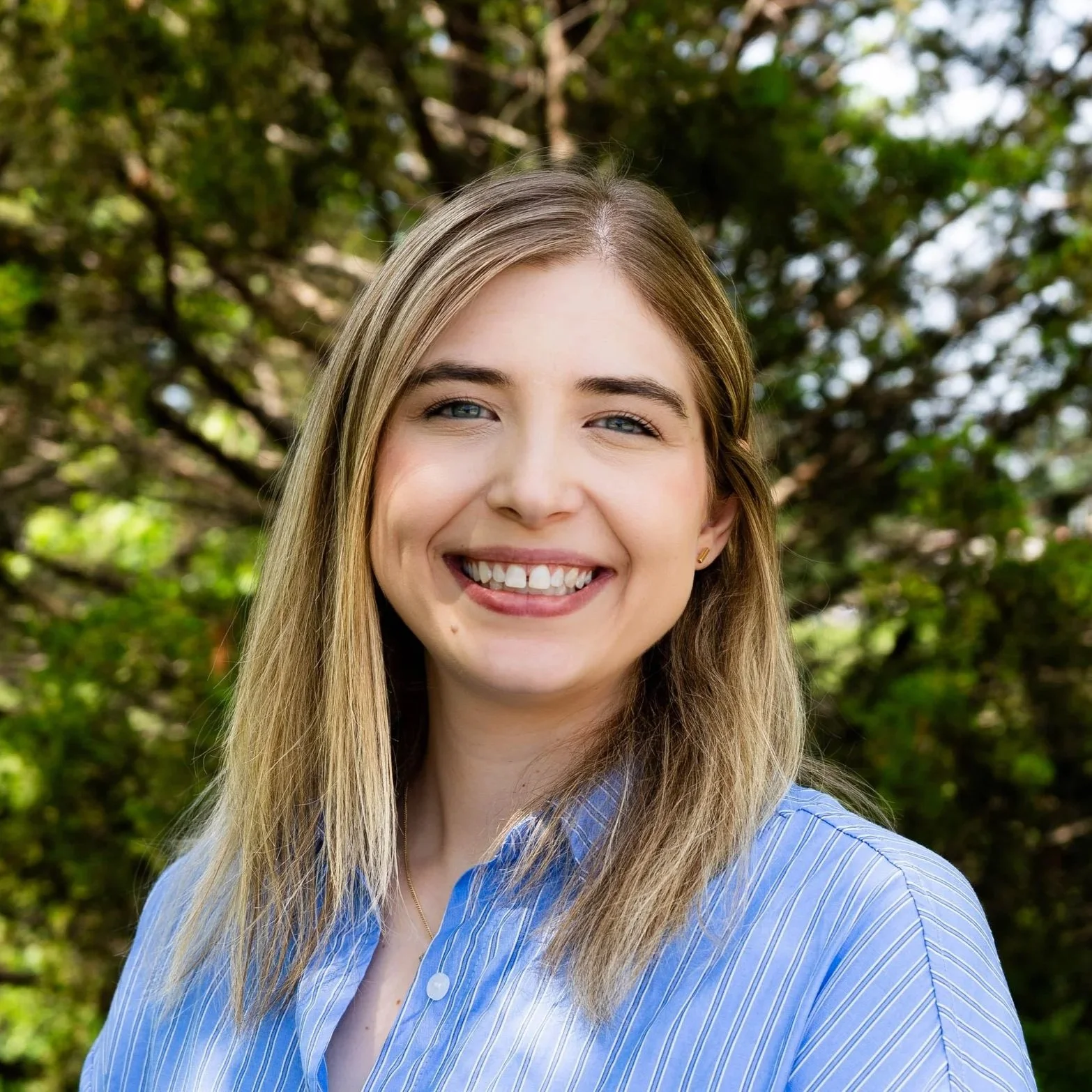 A young woman with long blonde hair, blue eyes, and a bright smile standing outdoors with trees in the background, wearing a blue and white striped shirt.