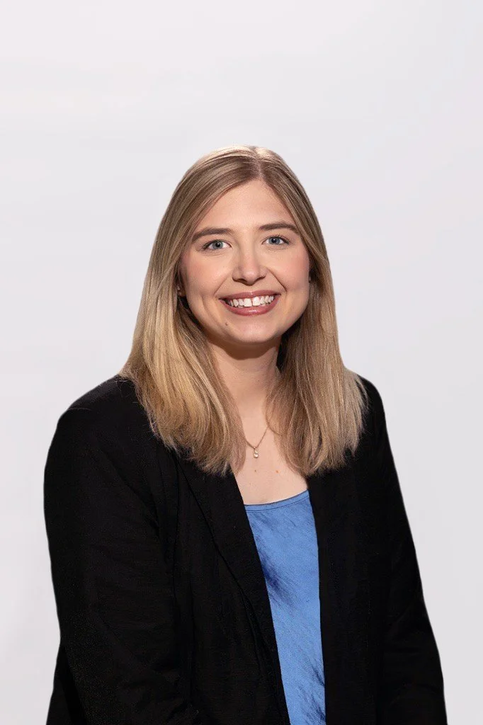 A young woman with shoulder-length blonde hair, wearing a black blazer and blue top, smiling against a plain white background.