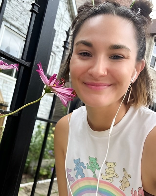 Young woman smiling outside, wearing a white sleeveless shirt with colorful animals and a rainbow, holding a pink flower, with earphones in her ears.