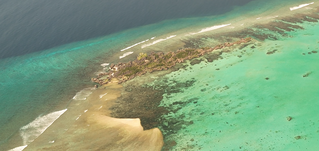 Aerial view of a small island surrounded by turquoise ocean waters, featuring a rocky coastline and patches of vegetation.
