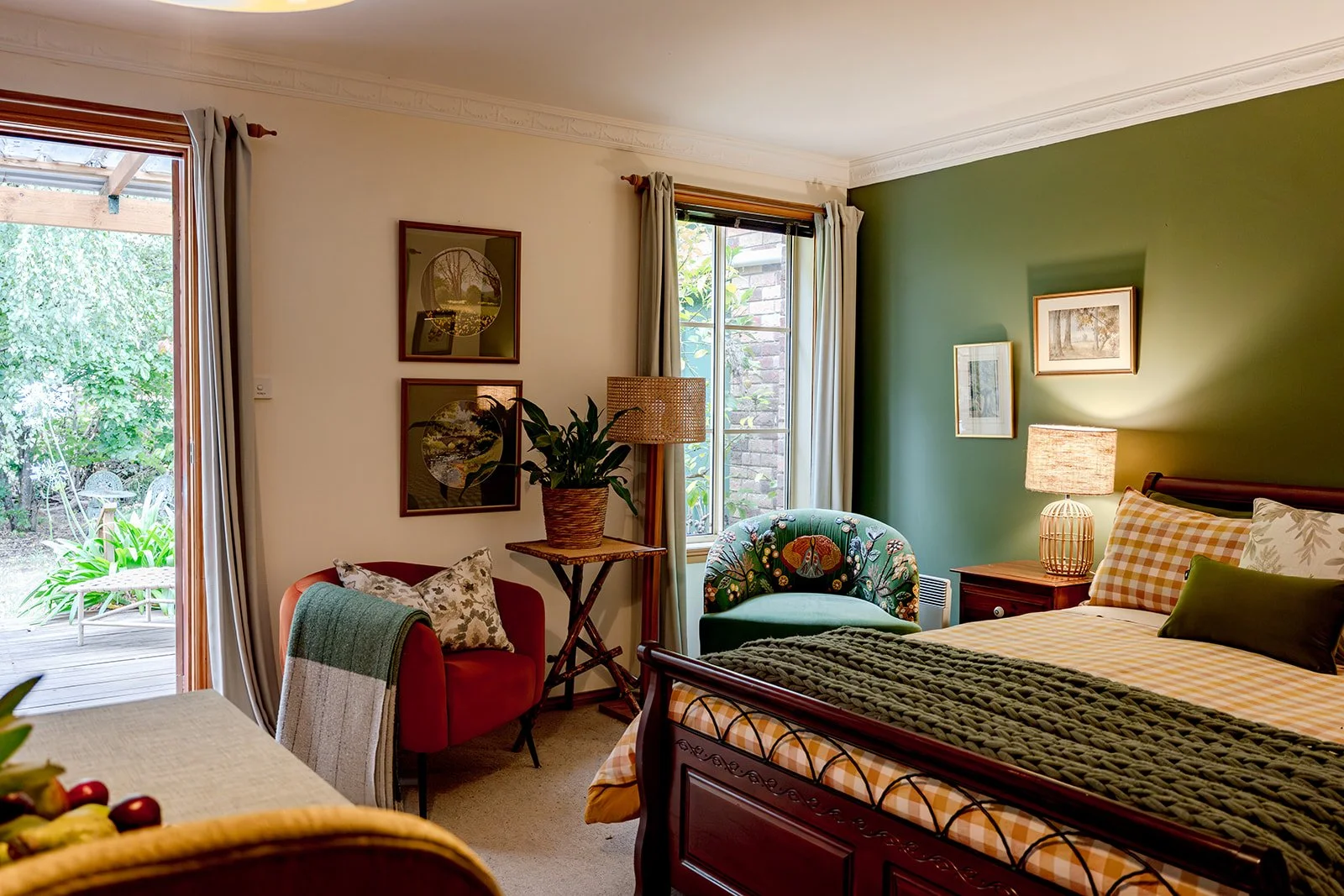 A cozy bedroom with a green accent wall, a wooden bed with striped and solid bedding, two bedside tables with lamps, a white armchair with floral cushions, and framed landscape paintings.
