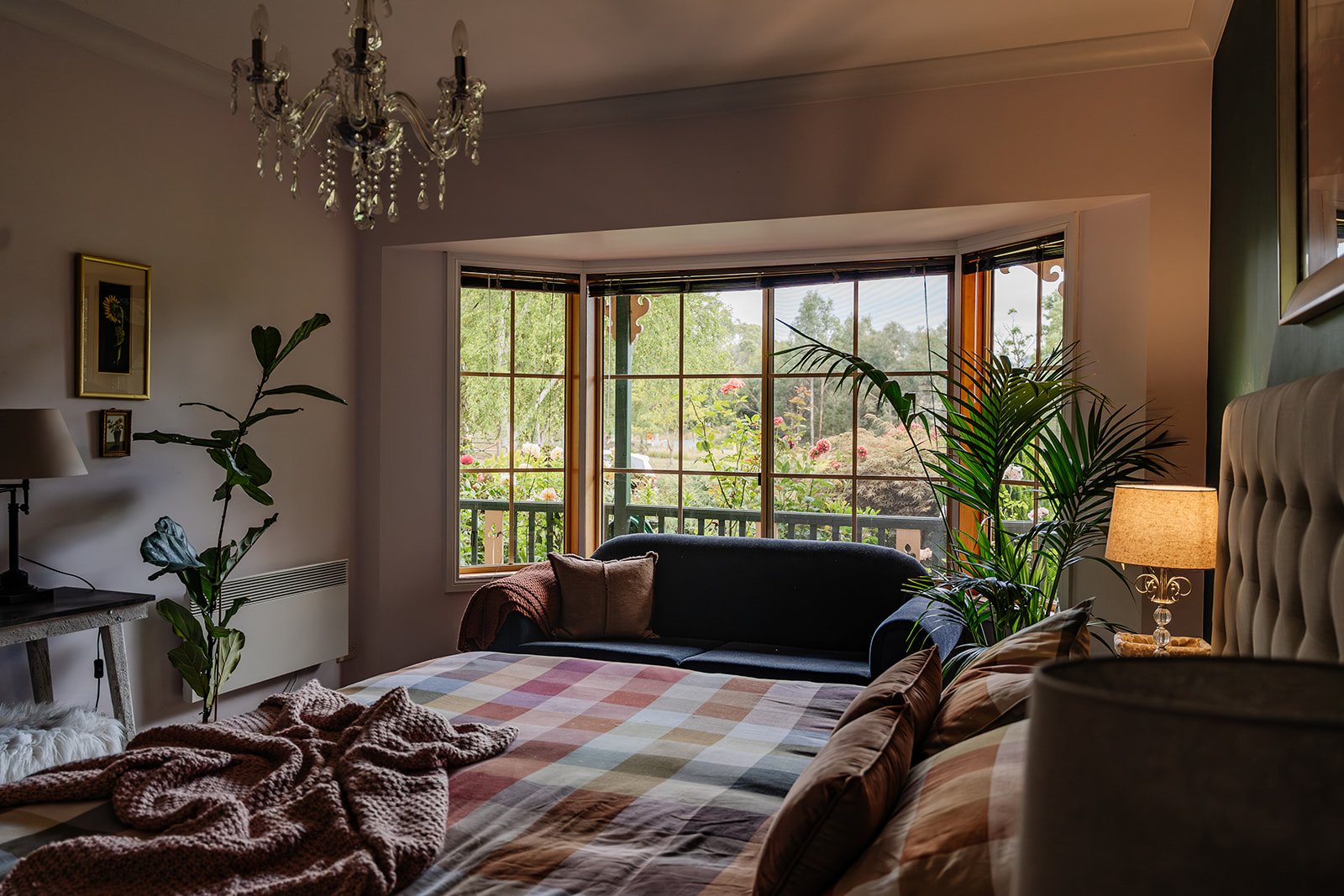 A neatly made bed with yellow and checkered pink and yellow bedding, topped with a chunky knitted throw, in a cozy bedroom with dark teal walls, white bedside tables with lamps, and a framed black and white photograph of Paris on the wall. A chandelier hangs from the ceiling.