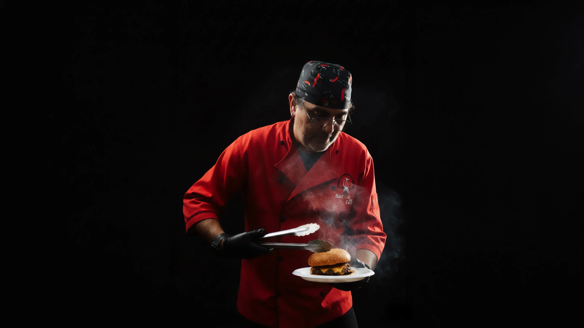 Chef argentino en fondo negro vestido de filipina roja preparando una hamburguesa con luz dramática