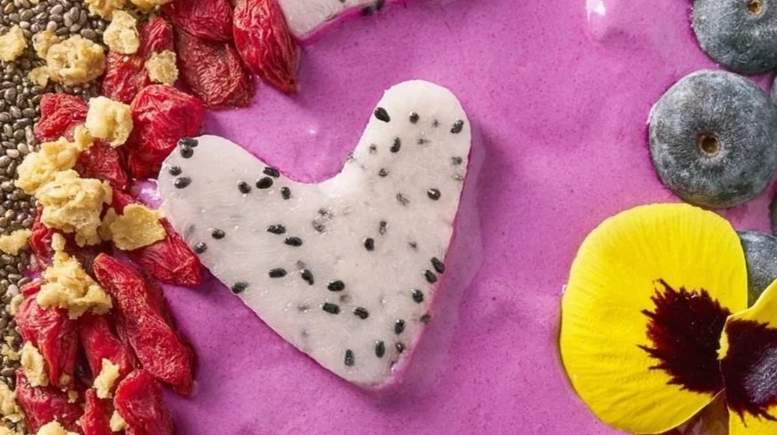 Heart-shaped piece of white chocolate with black sesame seeds surrounded by dried berries, a yellow flower, and blueberries on pink background.