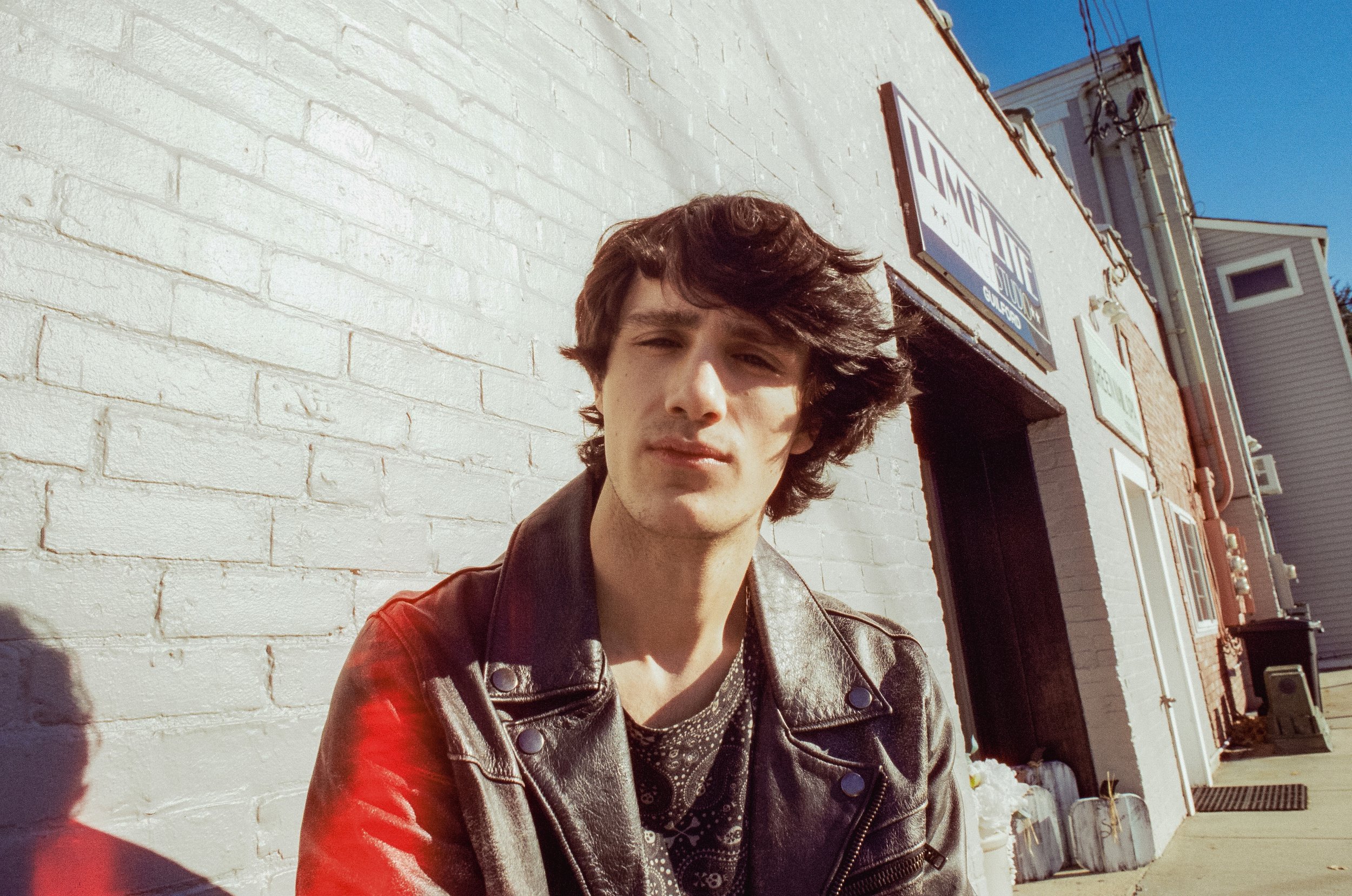 Young man with messy brown hair wearing a leather jacket, standing outdoors against a white brick wall, with sunlight and blue sky visible.