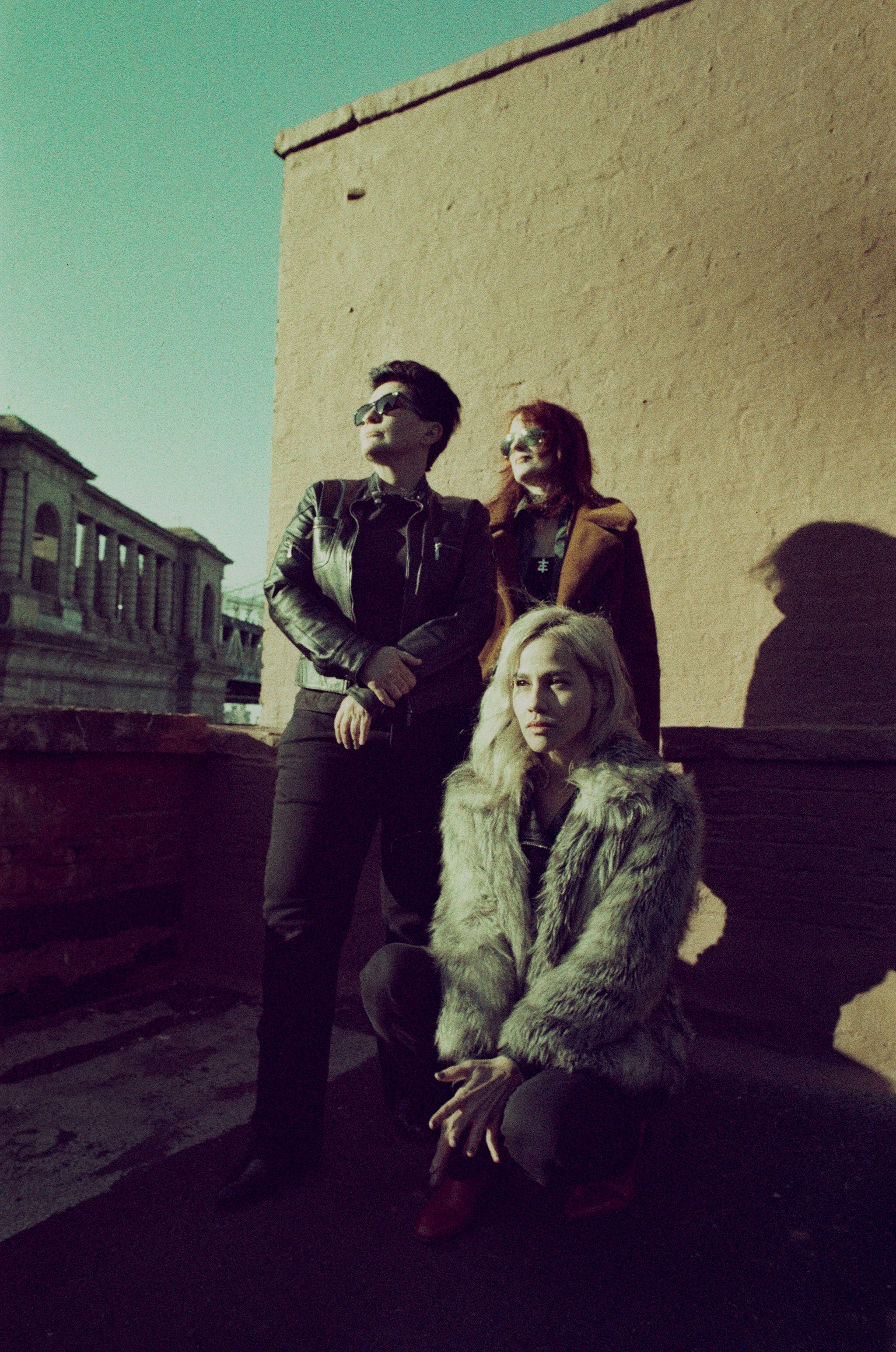 Three women posing on a rooftop with a beige wall in the background and a building in the distance, all wearing stylish clothing and sunglasses.