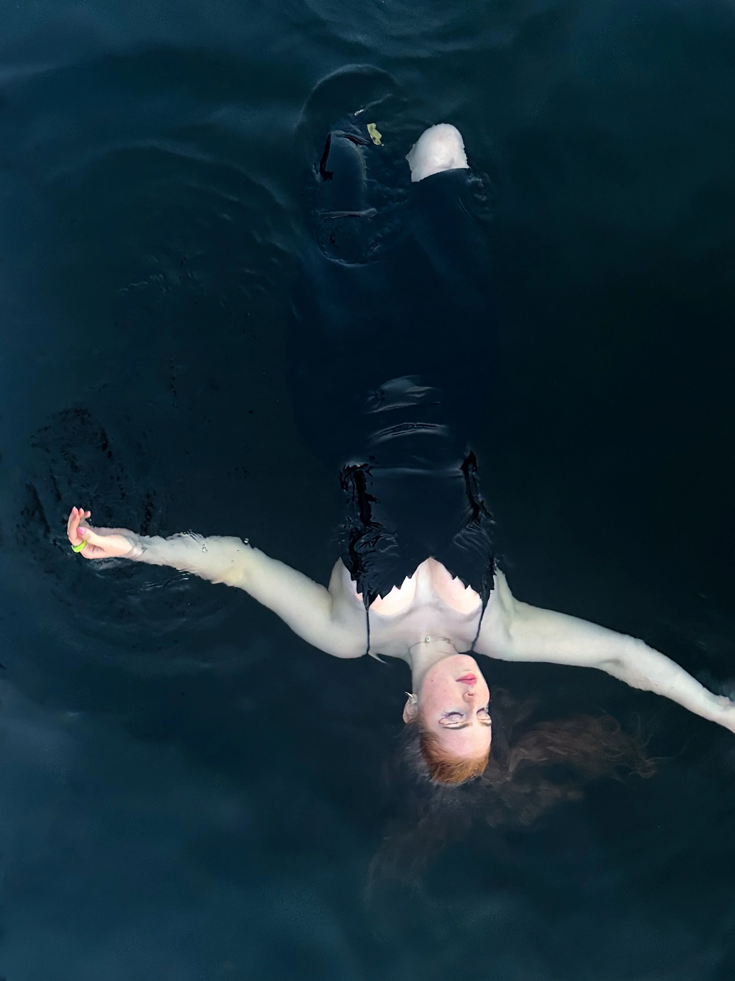 Person with red hair and fair skin lying on their back in dark water with arms and legs extended, wearing a black dress.