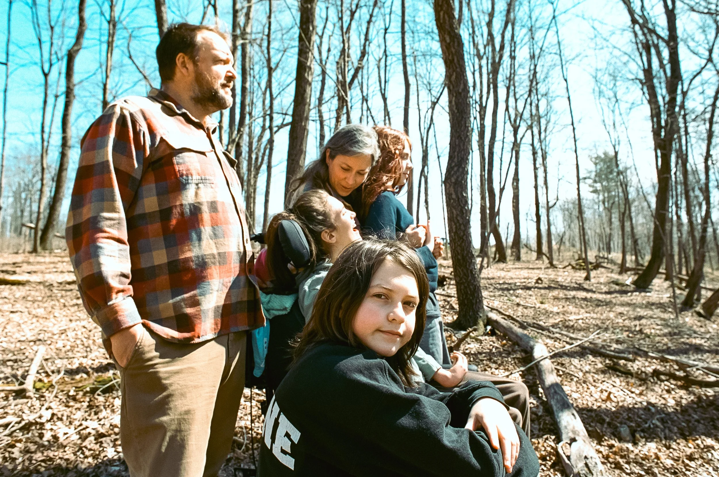 A group of six people, including children and adults, standing and sitting in a wooded area during spring, some sitting on fallen branches, enjoying a sunny day in the forest.
