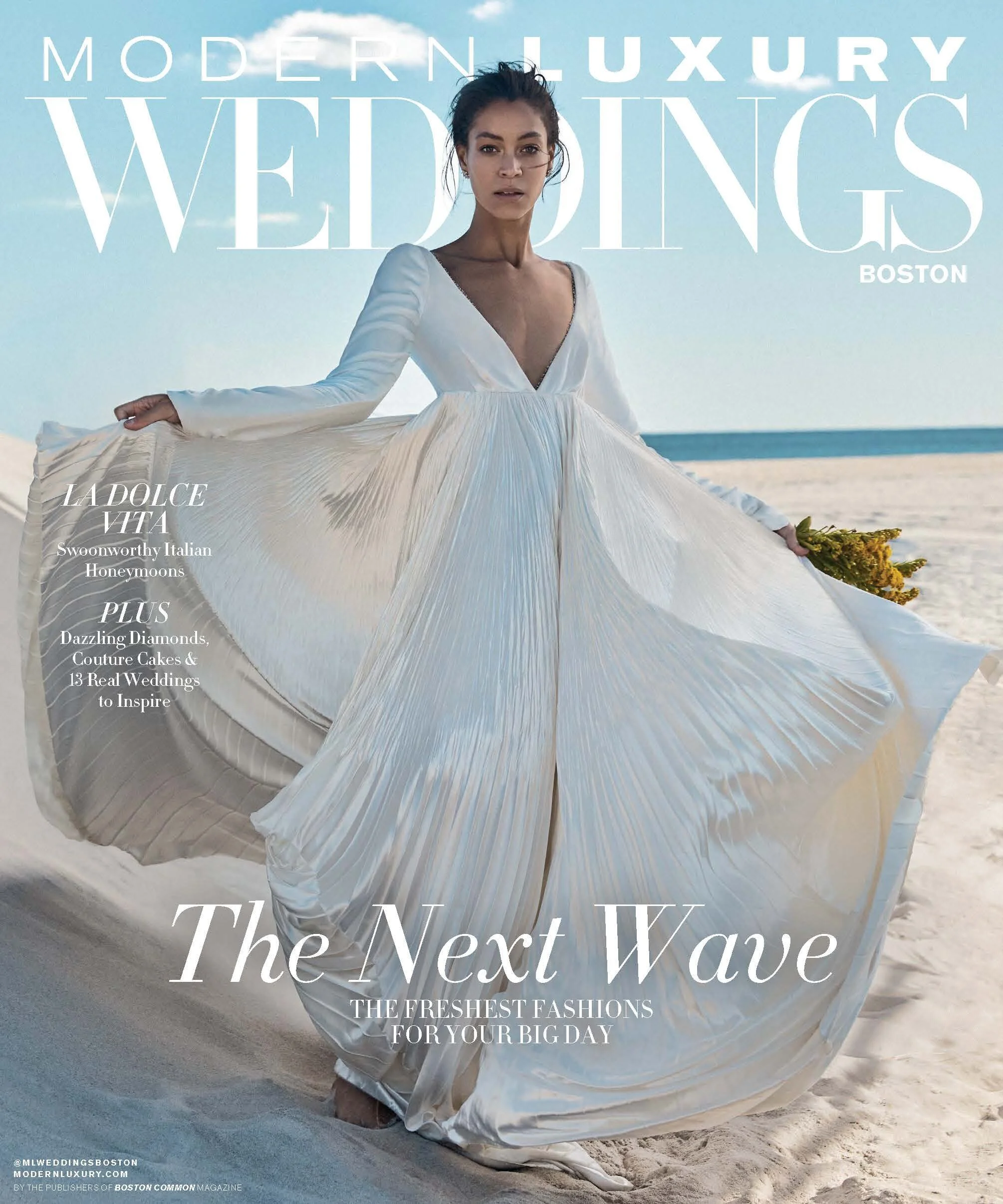 Woman in a flowing white dress standing on a sandy beach with an ocean background, holding the dress out to the sides.