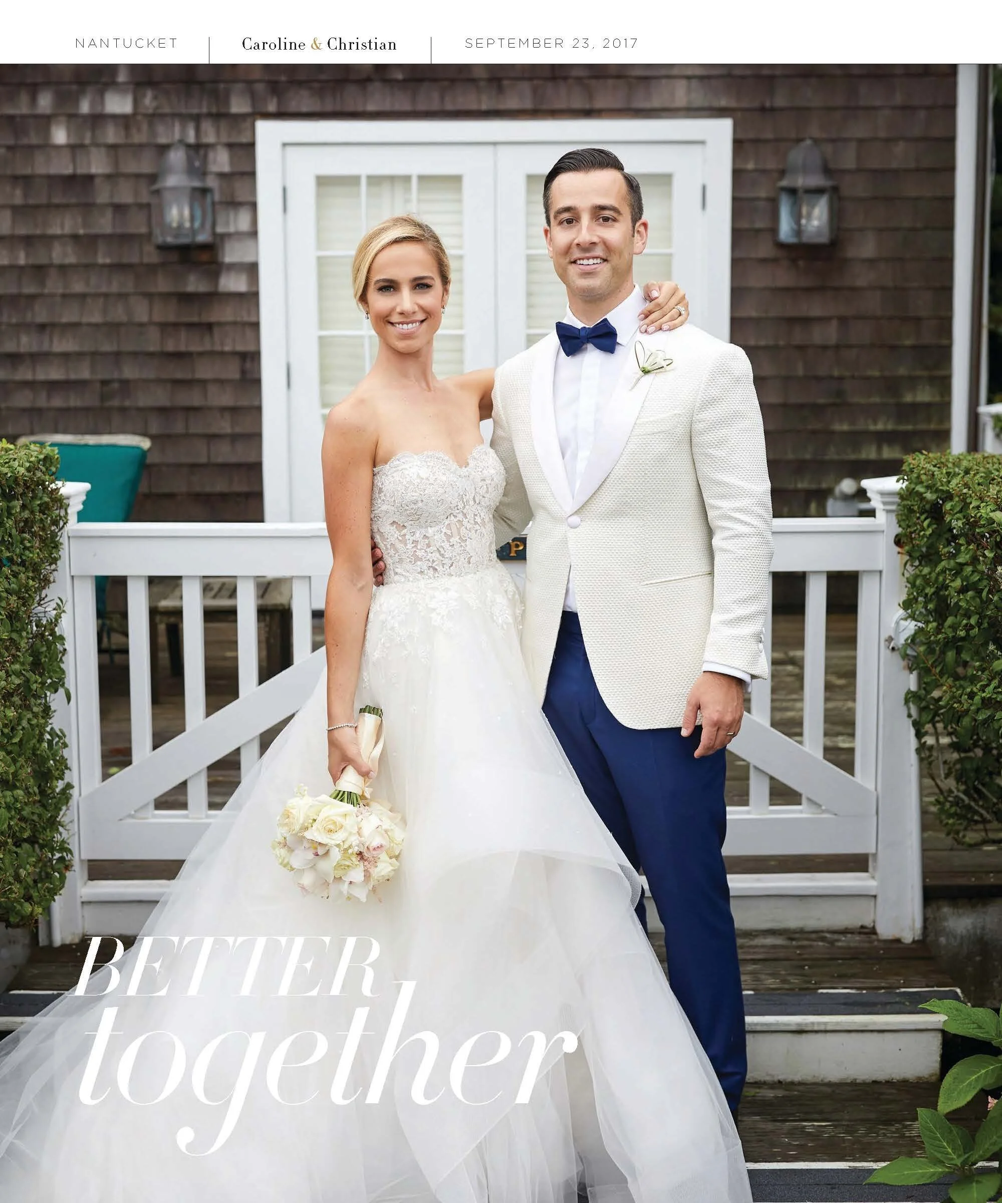 A newlywed couple smiling and standing outdoors on a wooden deck in front of a white fence and dark wooden house. The bride is wearing a strapless lace wedding dress and holding a bouquet of white flowers. The groom is dressed in a cream blazer with 