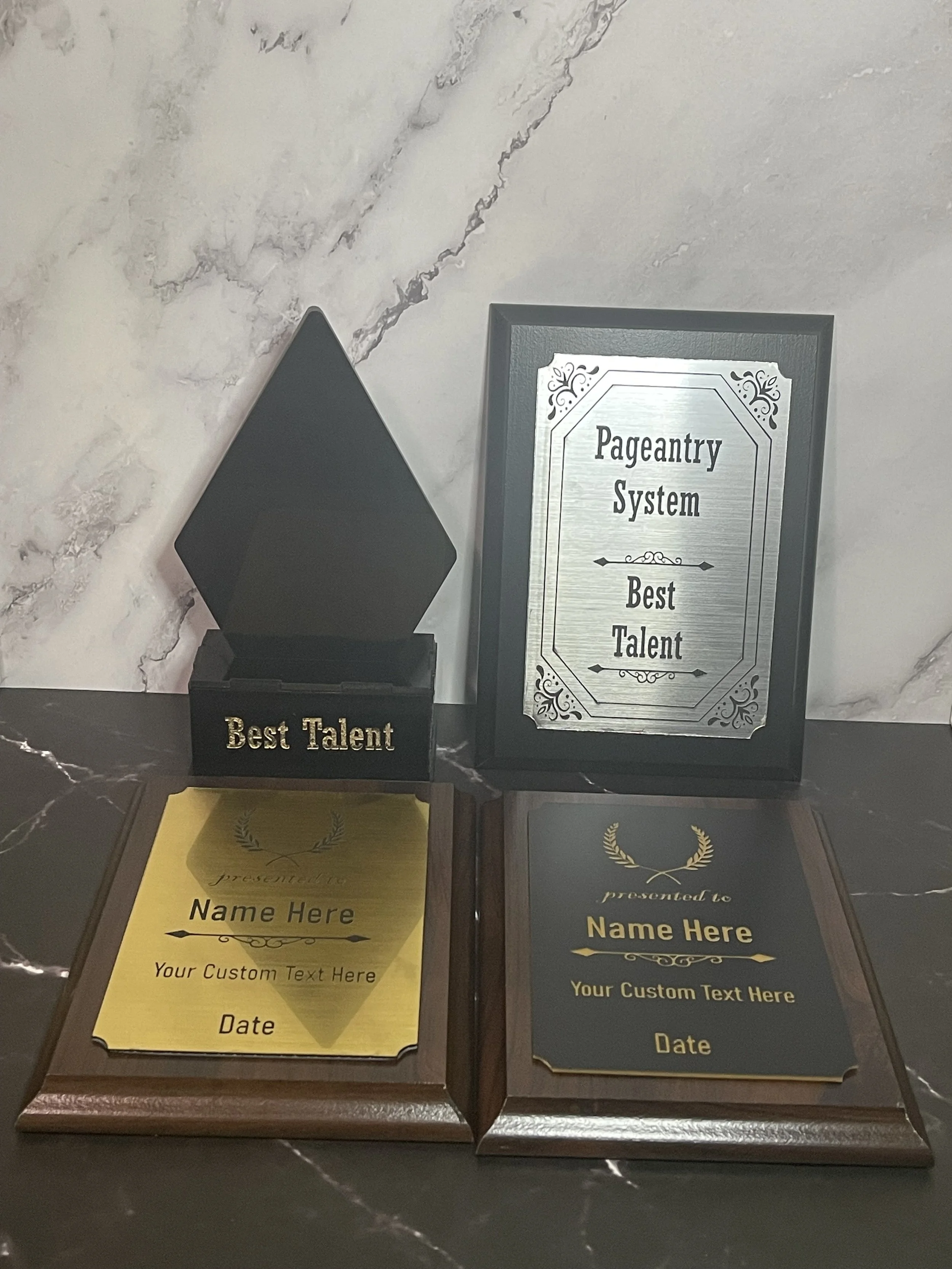 Award plaques and a sign for a pageantry system, including two certificates with customizable text, a box with a lid labeled 'Best Talent,' and a framed sign reading 'Pageantry System Best Talent' on a marble surface.
