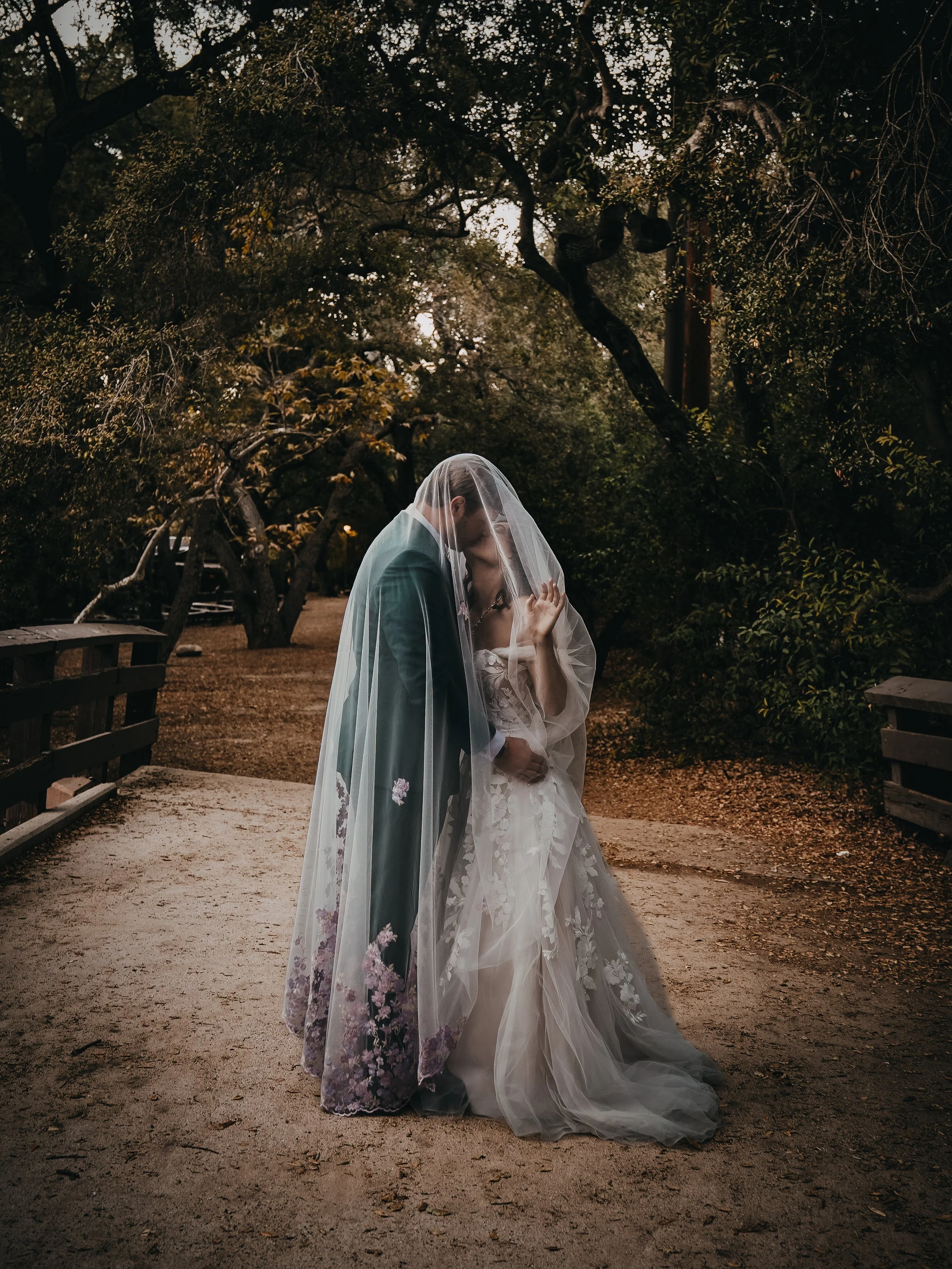 A bride and groom share a kiss under a wedding veil on a park bridge surrounded by trees.