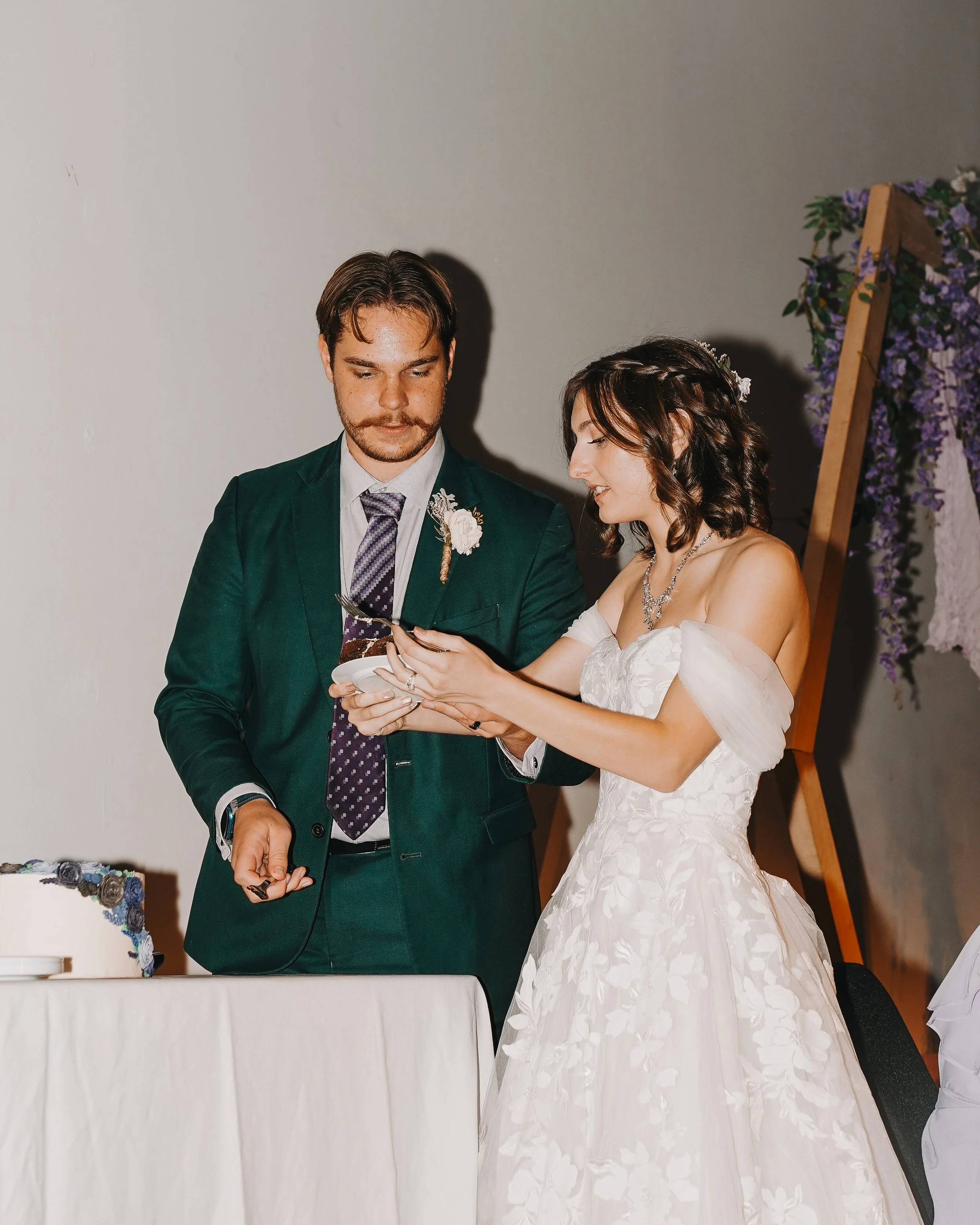 A bride in a white wedding dress and a groom in a dark green suit exchanging gifts during their wedding reception, standing by a table with a cake.