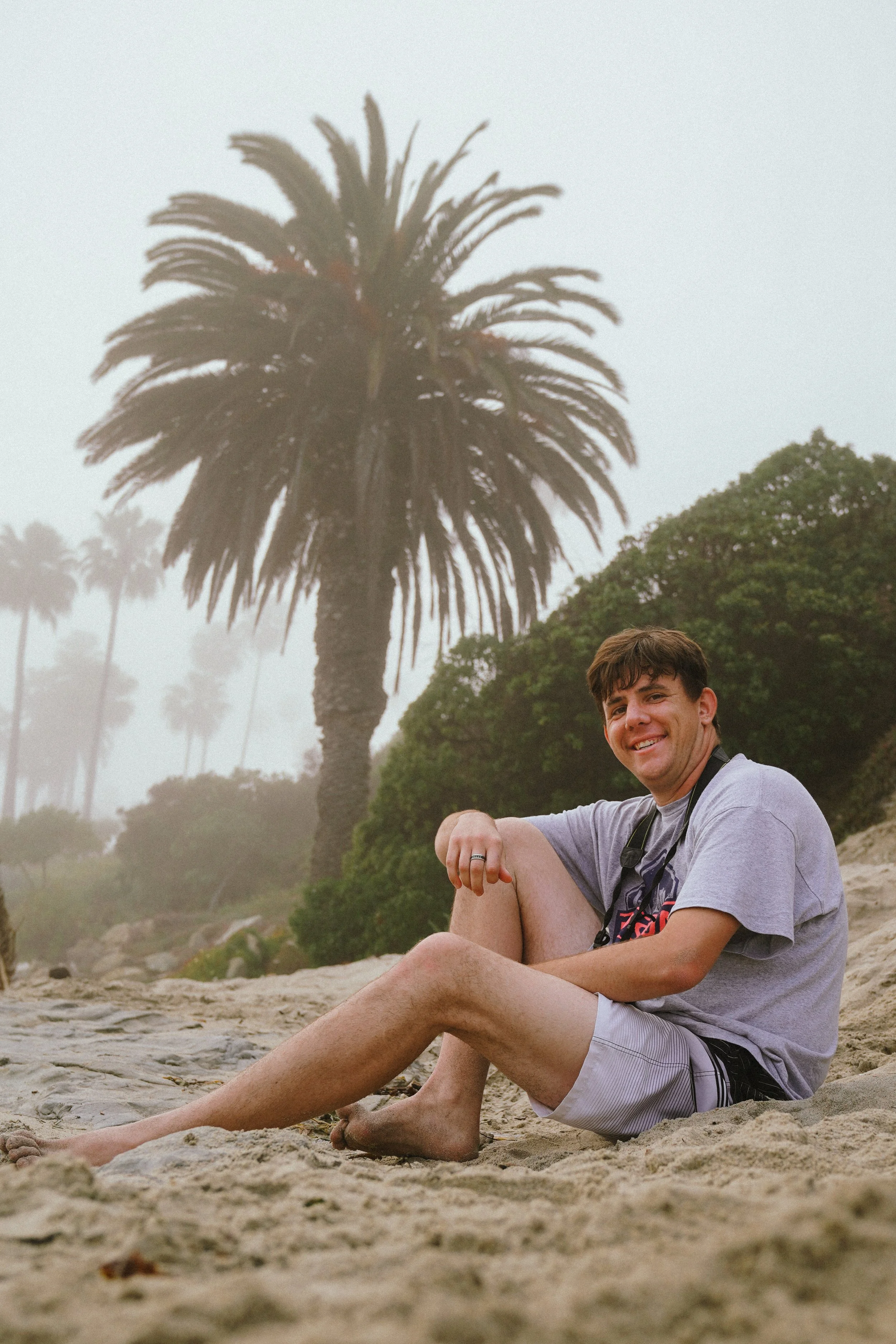 A smiling man sitting on sandy ground with palm trees and fog in the background.