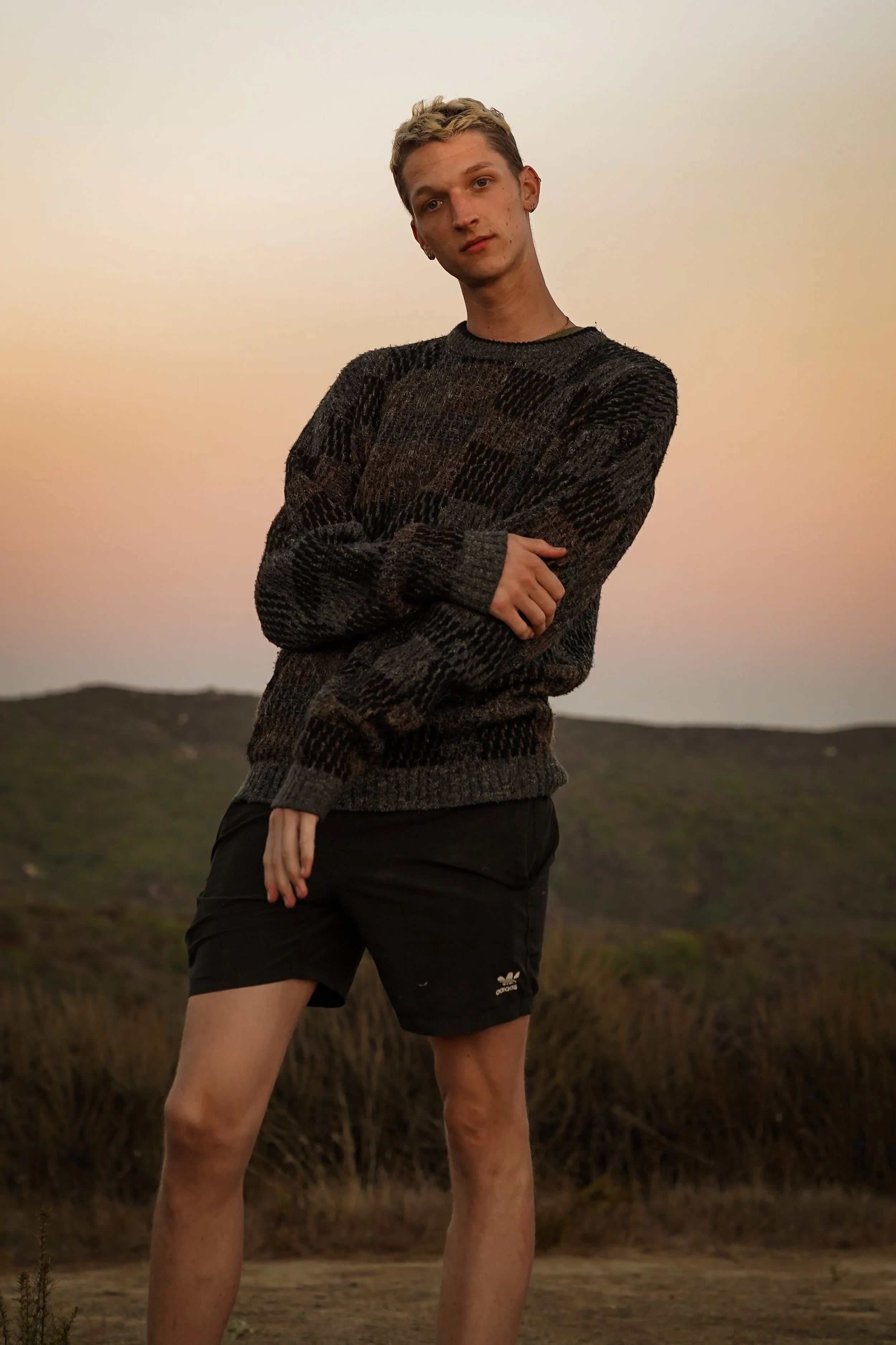 A young man standing outdoors at sunset, wearing a dark patterned sweater and black shorts, with hills and dry grass in the background.