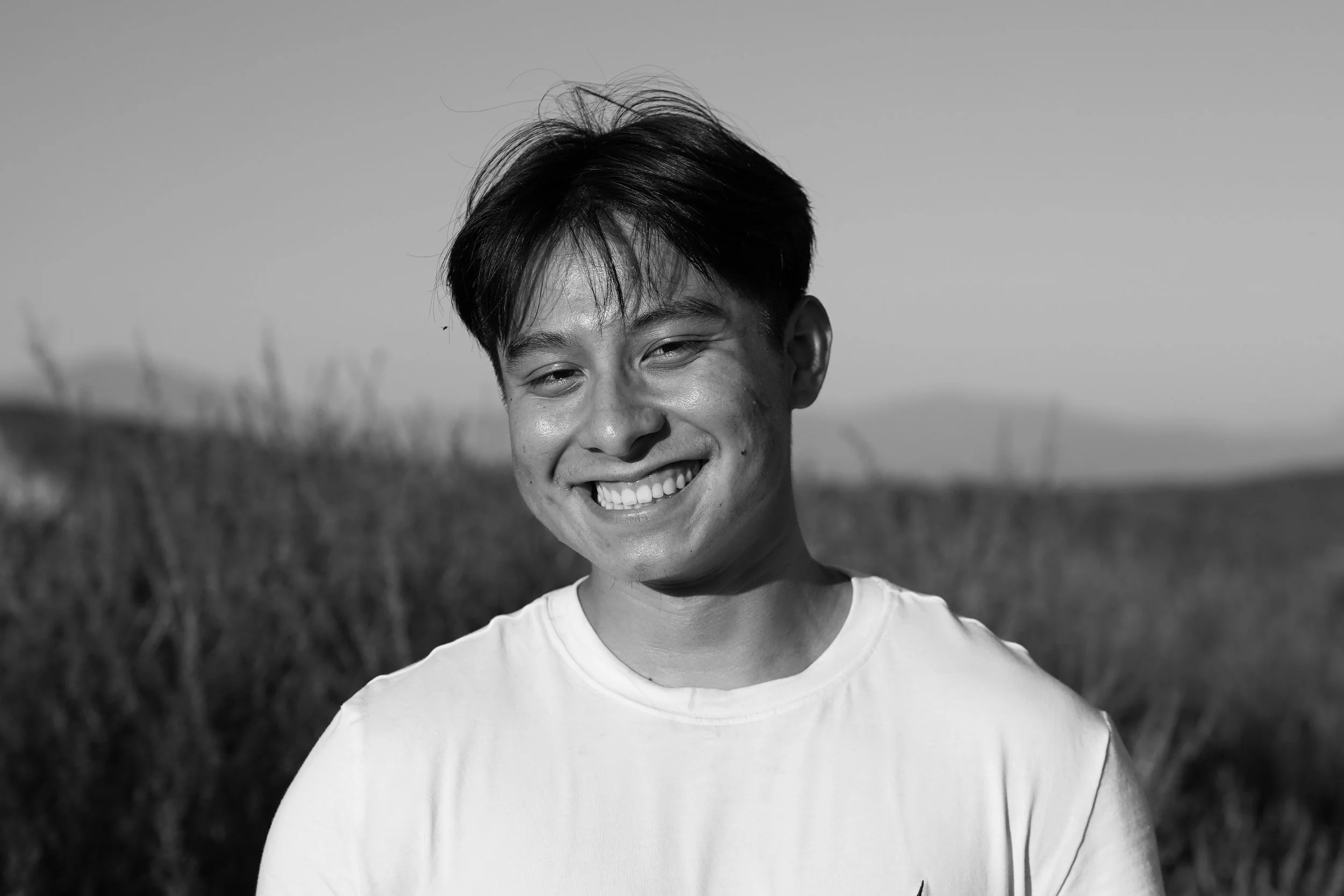 Black and white photo of a smiling young man with short dark hair, wearing a light-colored t-shirt, outdoors in a field with tall grass.