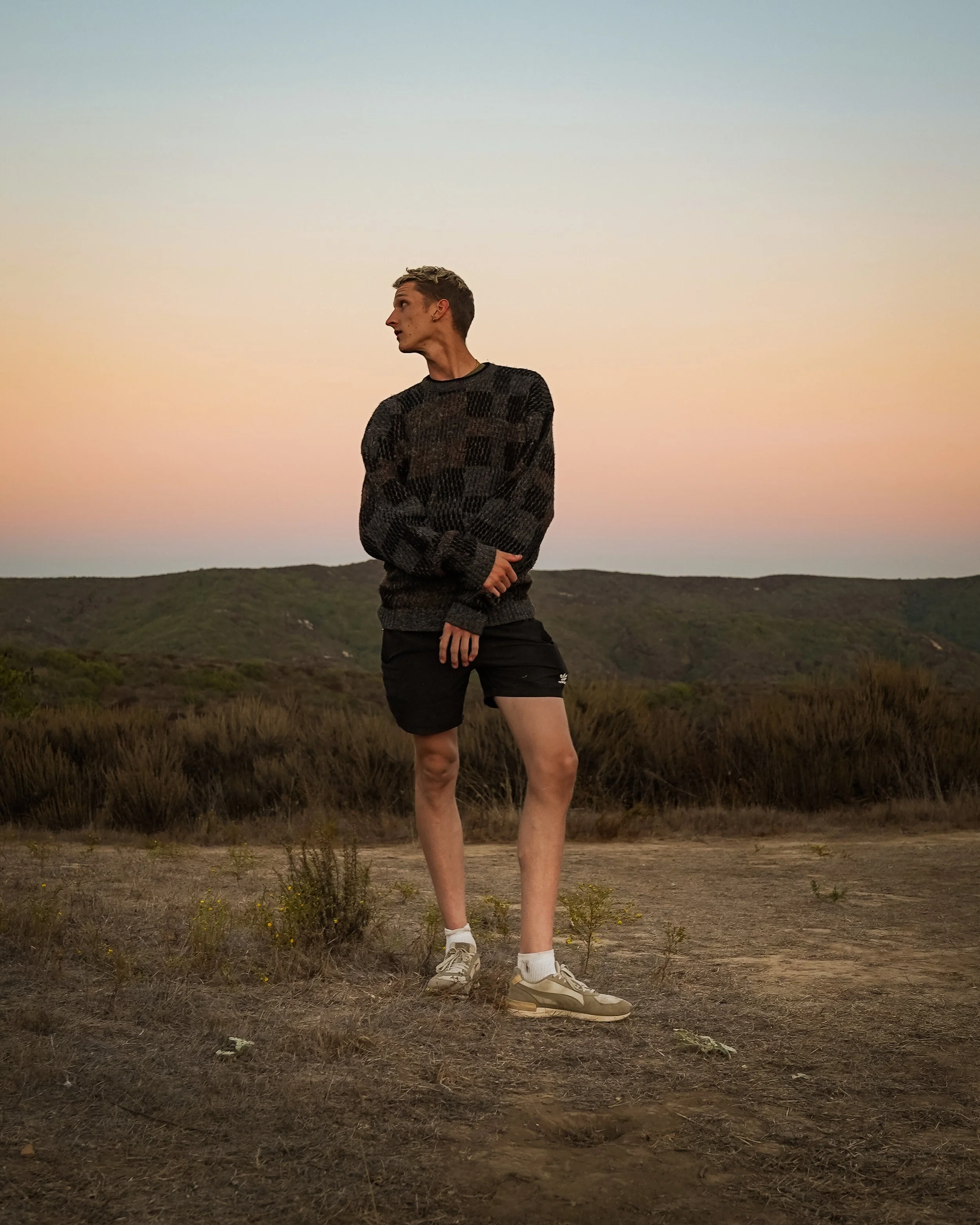 A young man standing outdoors at sunset, wearing a patterned sweater, black shorts, white socks, and sneakers, with a landscape of hills and bushes in the background.
