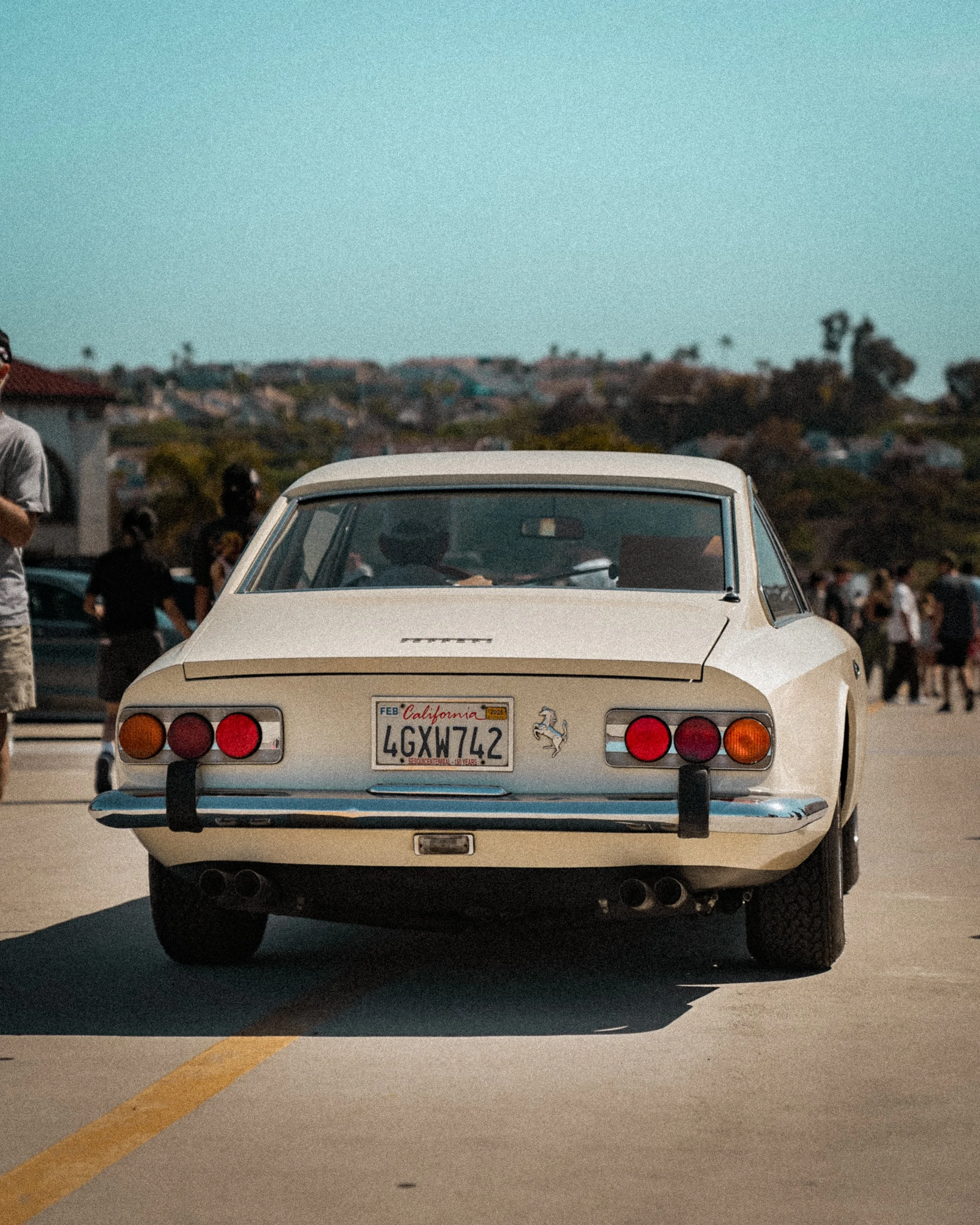 Back view of a vintage white Ferrari 365 GT4/BB car parked outdoors, with a California license plate and a Ferrari emblem on the trunk, surrounded by people and other vehicles.