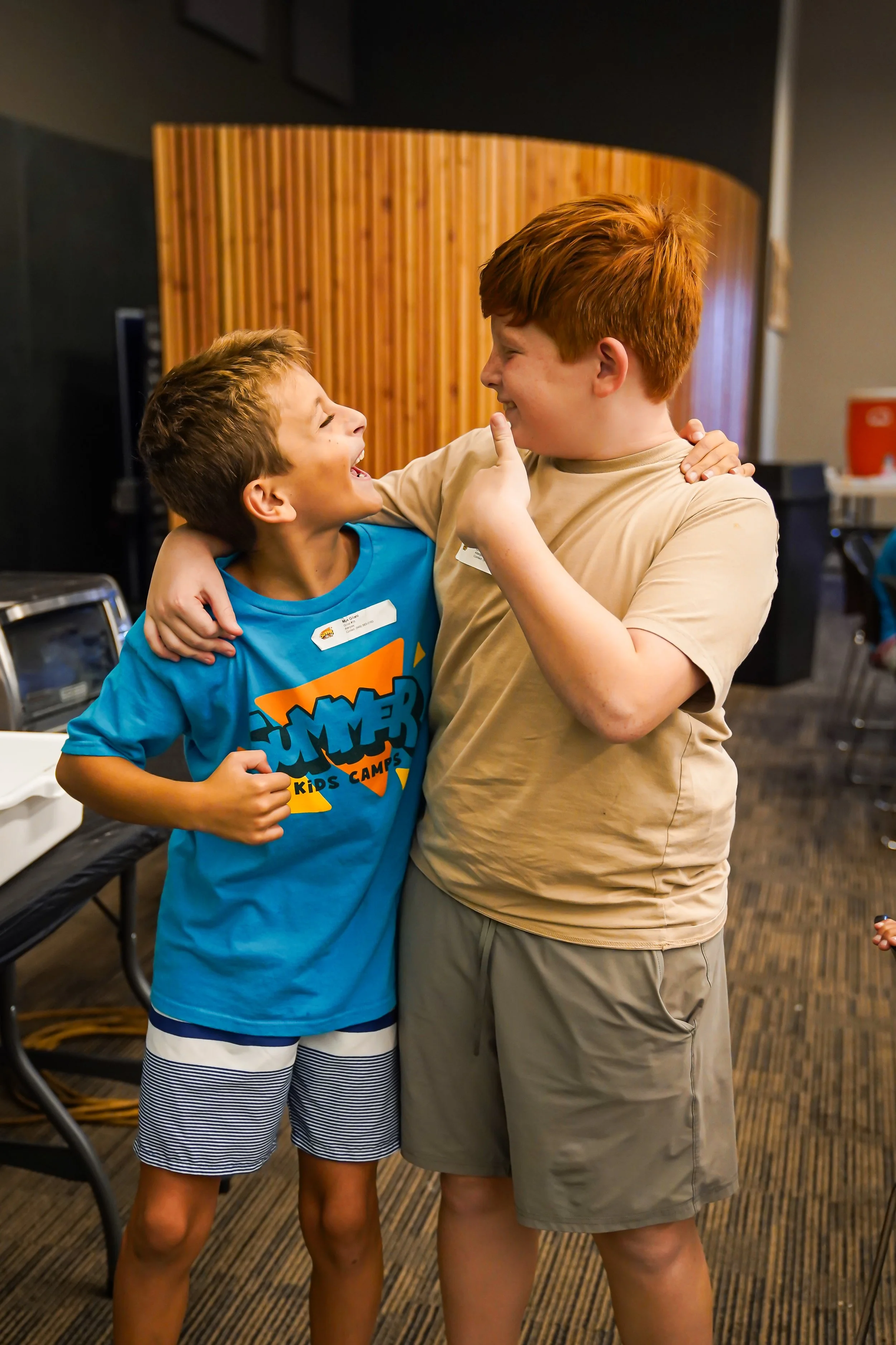 Two boys smiling and hugging each other indoors, one in a blue t-shirt and the other in a beige t-shirt.