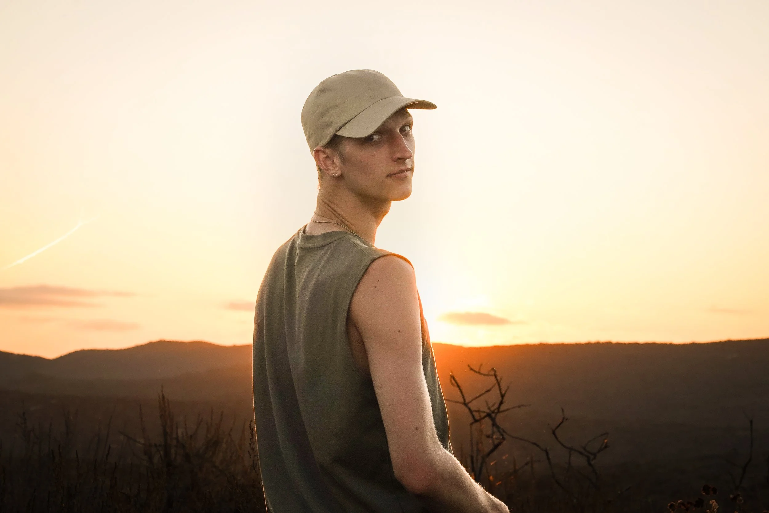 A young person with short hair, wearing a beige cap and sleeveless shirt, standing outdoors during sunset or sunrise in a hilly landscape.