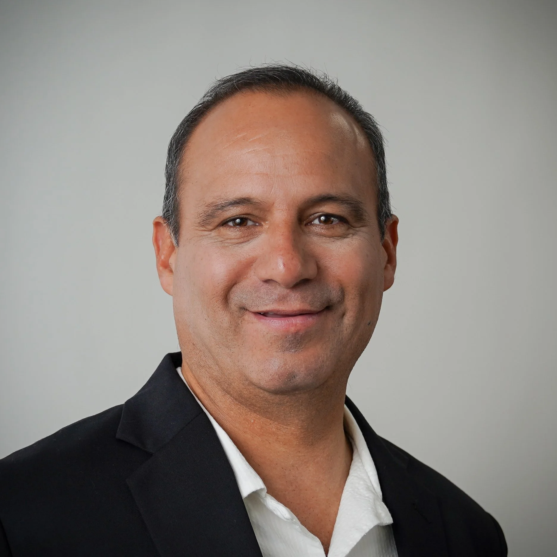 A woman with dark hair in a sleeveless cream-colored dress with embroidered flowers, standing outdoors among greenery and trees, with sunlight illuminating her face.