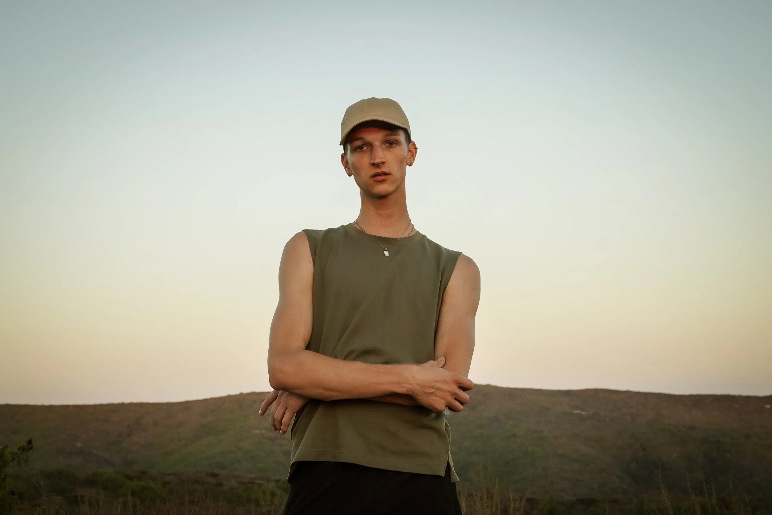 A young person standing outdoors on a hill at sunset, wearing a beige cap, a sleeveless olive green shirt, and a necklace, with arms crossed and a serious expression.