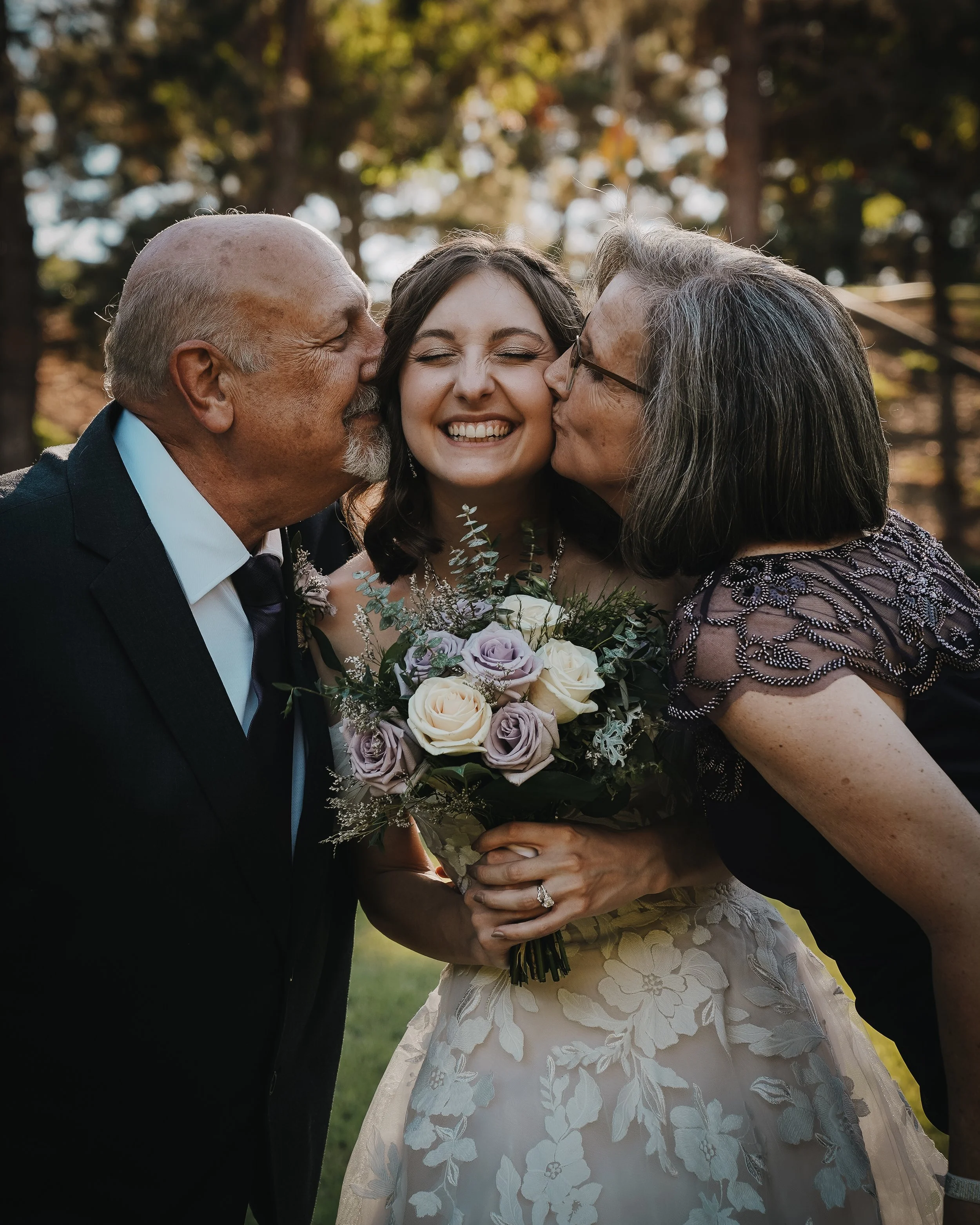 A young woman in a wedding dress holding a bouquet of flowers, smiling happily, being kissed on both cheeks by an older man and woman, likely her parents, outdoors during daytime.