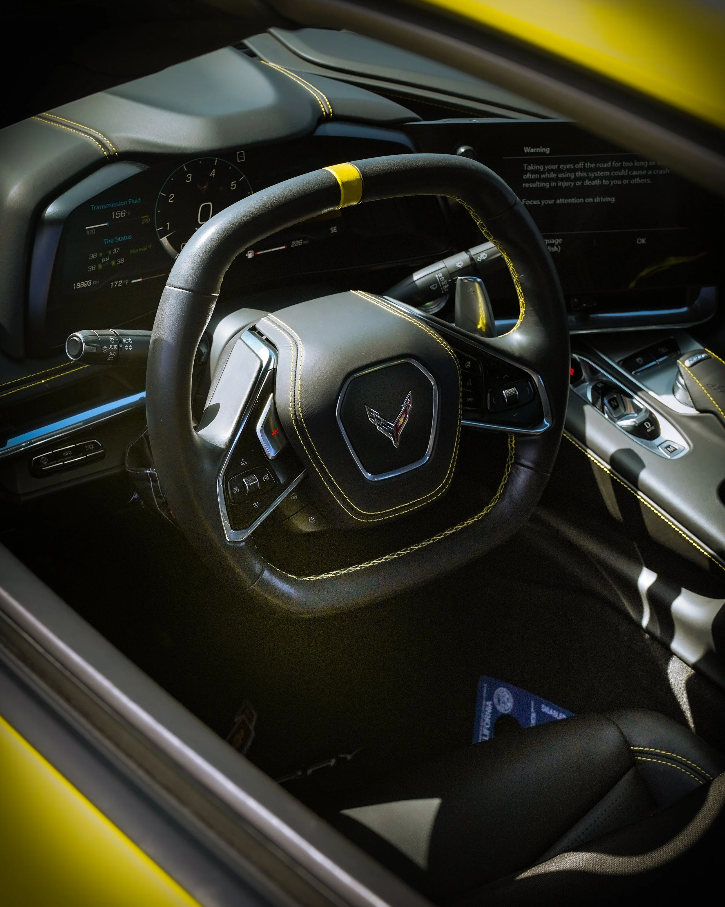 Interior of a Chevrolet Corvette sports car with a black leather steering wheel featuring yellow stitching and a Chevrolet emblem in the center, dashboard with digital displays, and yellow accents.
