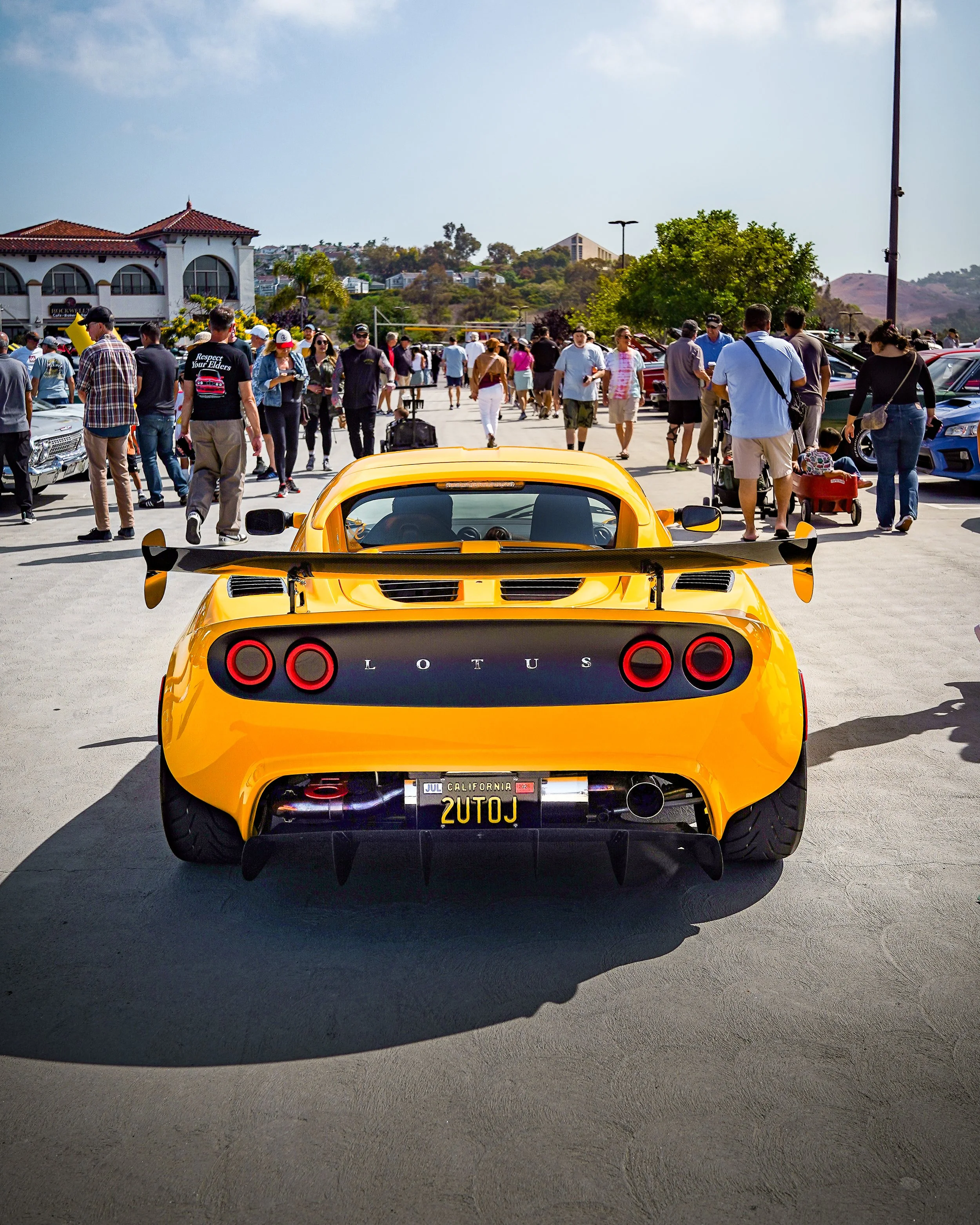 A yellow Lotus sports car parked outdoors at a car show, with a crowd of people walking in the background.