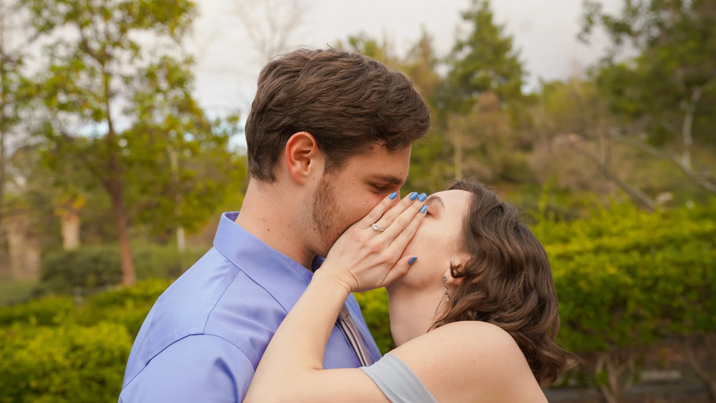 A couple kissing outdoors, with the woman touching the man's face. The woman has wavy brown hair and is wearing earrings, while the man has short brown hair and is dressed in a light blue button-up shirt. They are standing in a park with green trees 