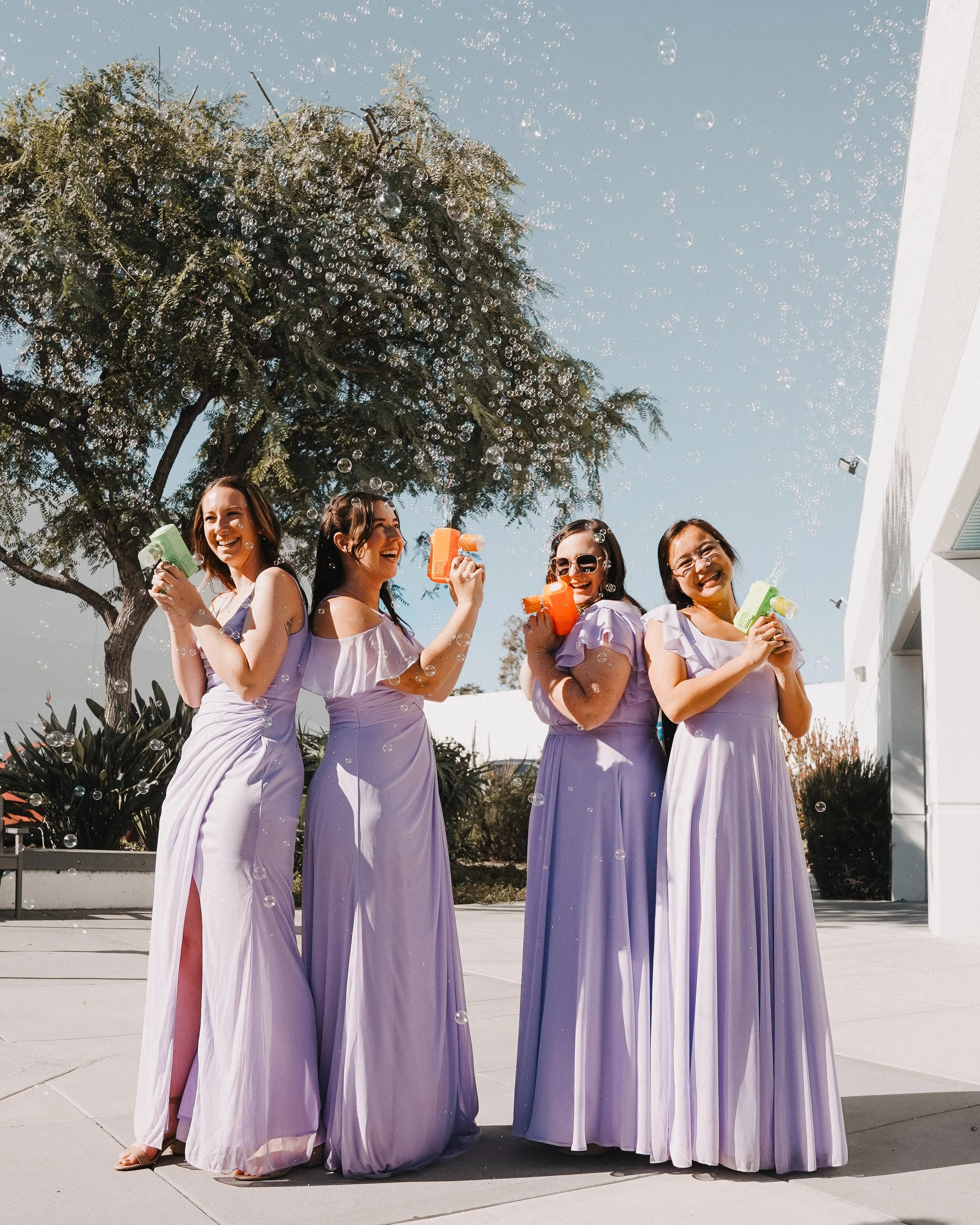 Bride smiling at groom as they walk outside, surrounded by friends and family. The bride wears a strapless white wedding gown and holds a bouquet, while the groom wears a dark suit with a boutonnière. People are celebrating and blowing bubbles in the background on a sunny day with trees and parked cars.