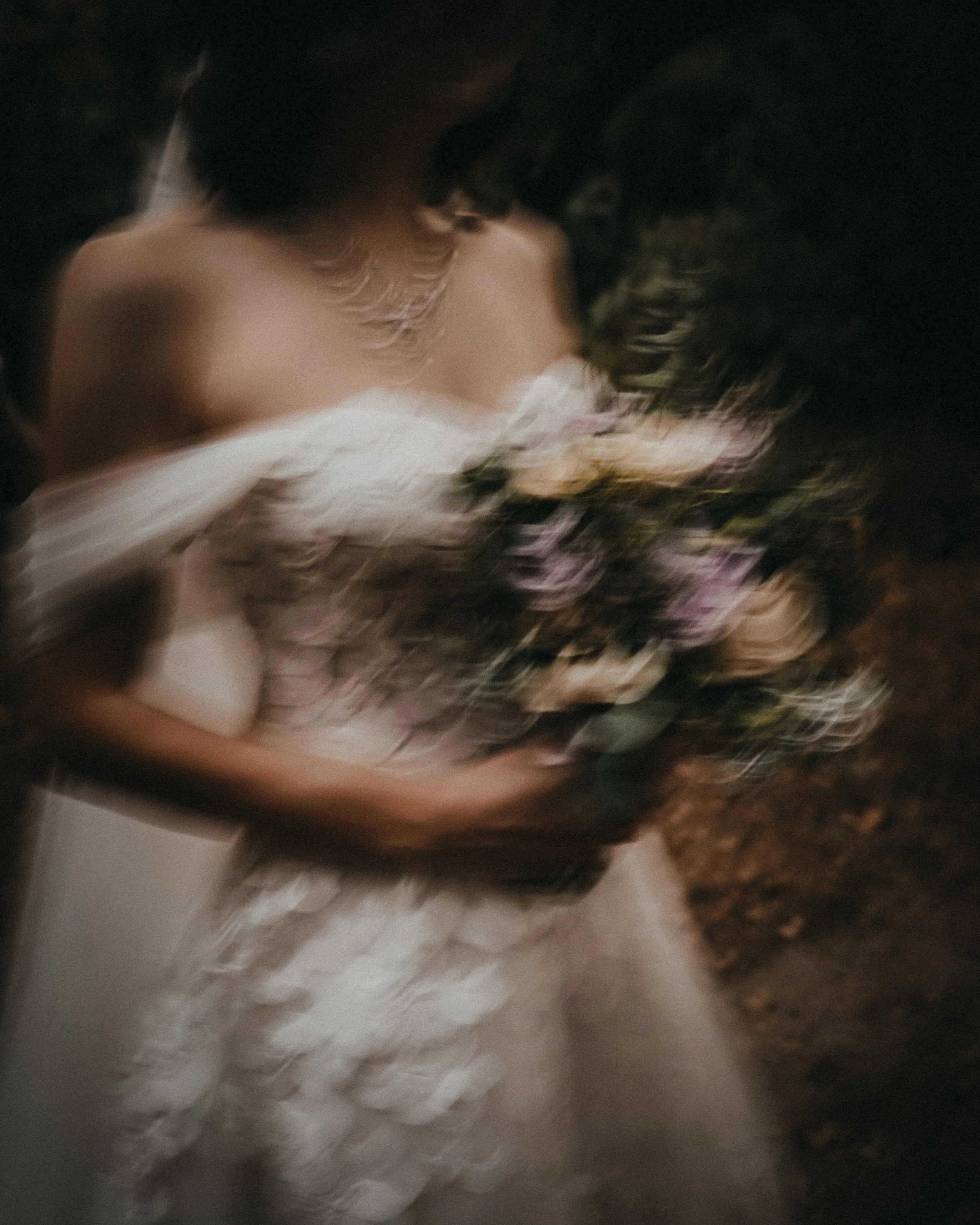 A blurred photo of a person holding a bouquet of flowers, possibly at a wedding, with a dark background.