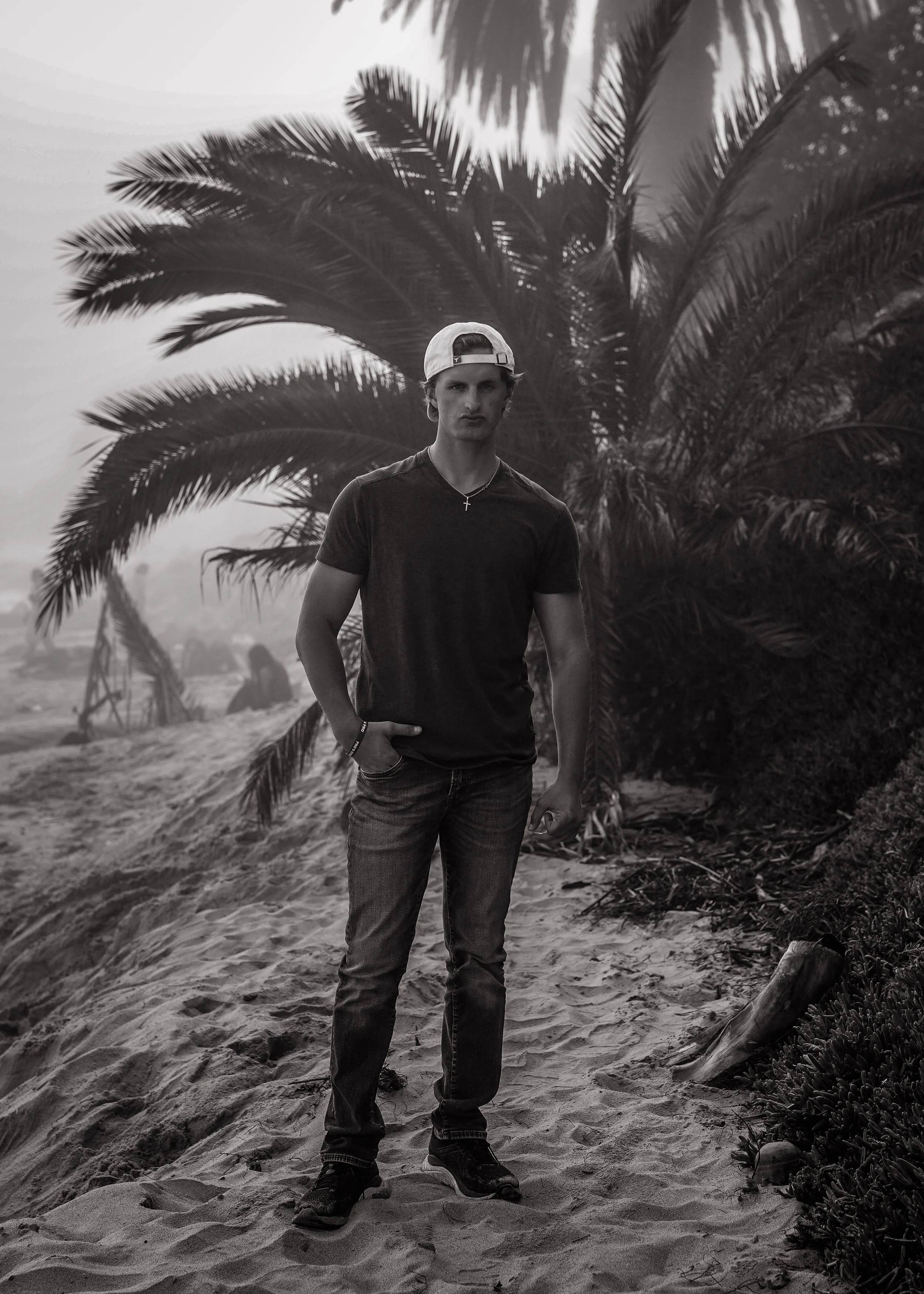 Young man standing on sandy ground near palm trees, wearing a backwards cap, dark t-shirt, jeans, and sneakers, in a natural outdoor setting.