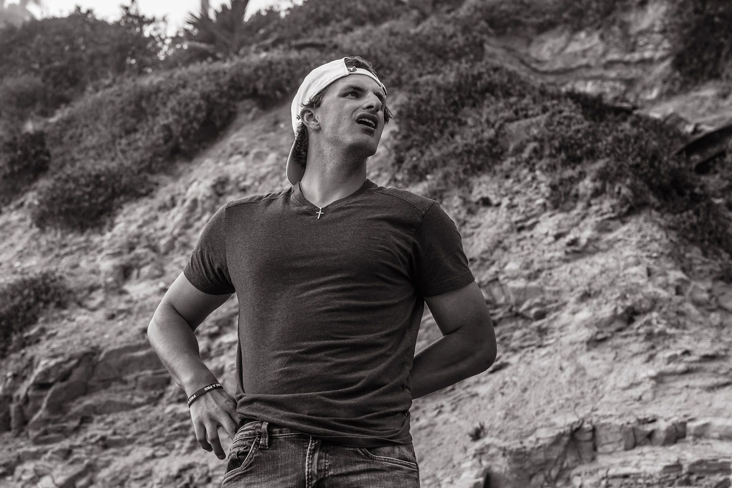 A young man wearing a backwards cap, a T-shirt, and jeans standing outdoors with a rocky hillside in the background, looking up with a thoughtful expression.