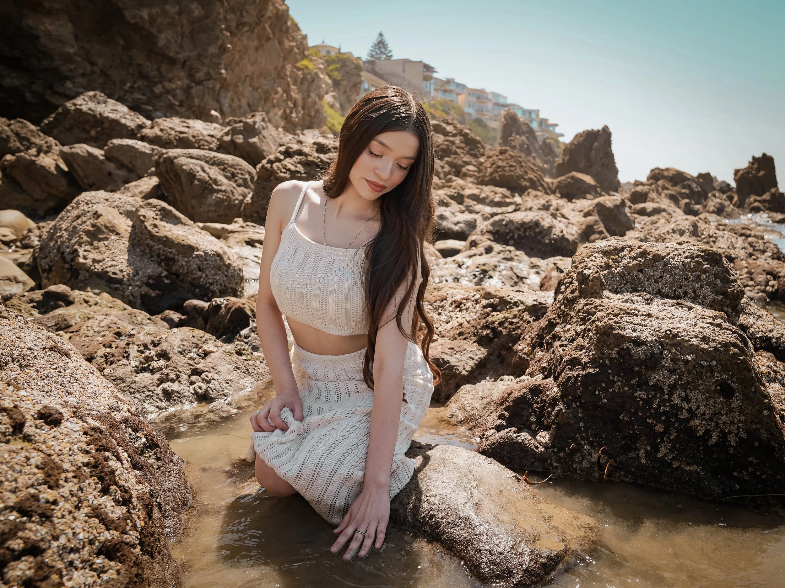 A woman with long dark hair kneels on rocks in shallow water at a rocky beach with houses on a hillside in the background.