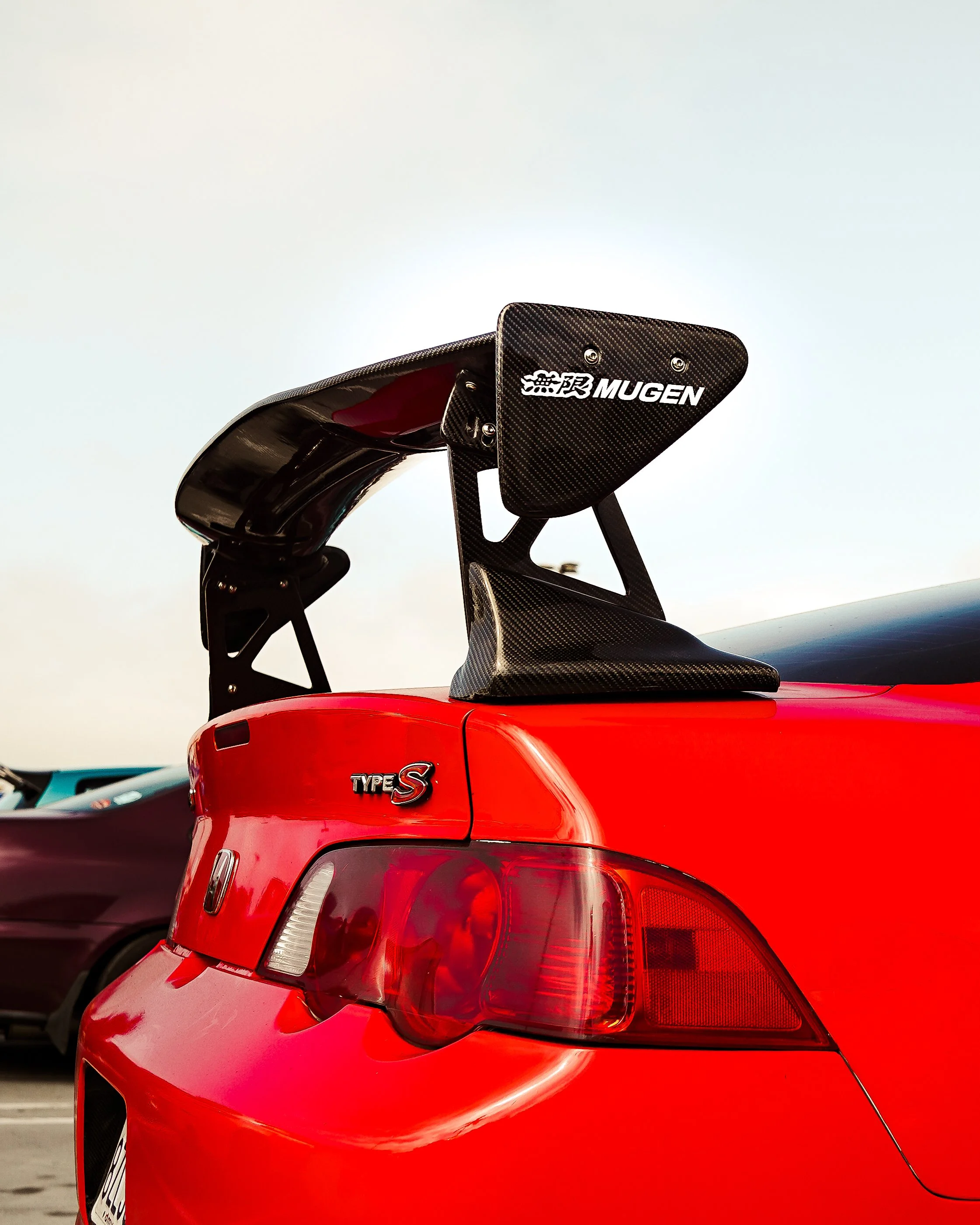 Close-up of the rear of a red sports car with a black wing spoiler labeled 'Mugen' and 'Type S' badge.