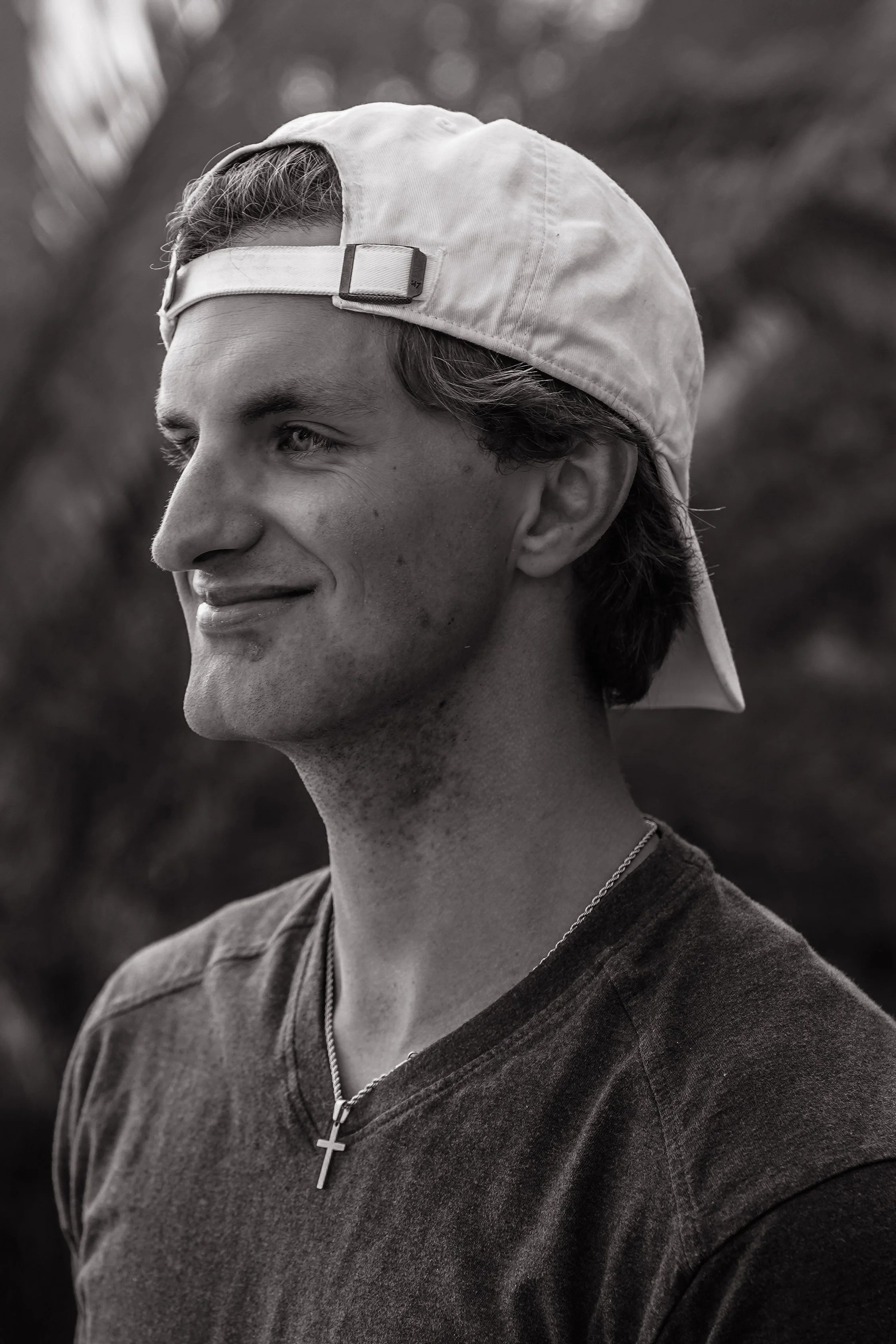 A young man with light skin, wearing a backward baseball cap, a cross necklace, and a dark T-shirt, smiling gently outdoors with trees in the background.