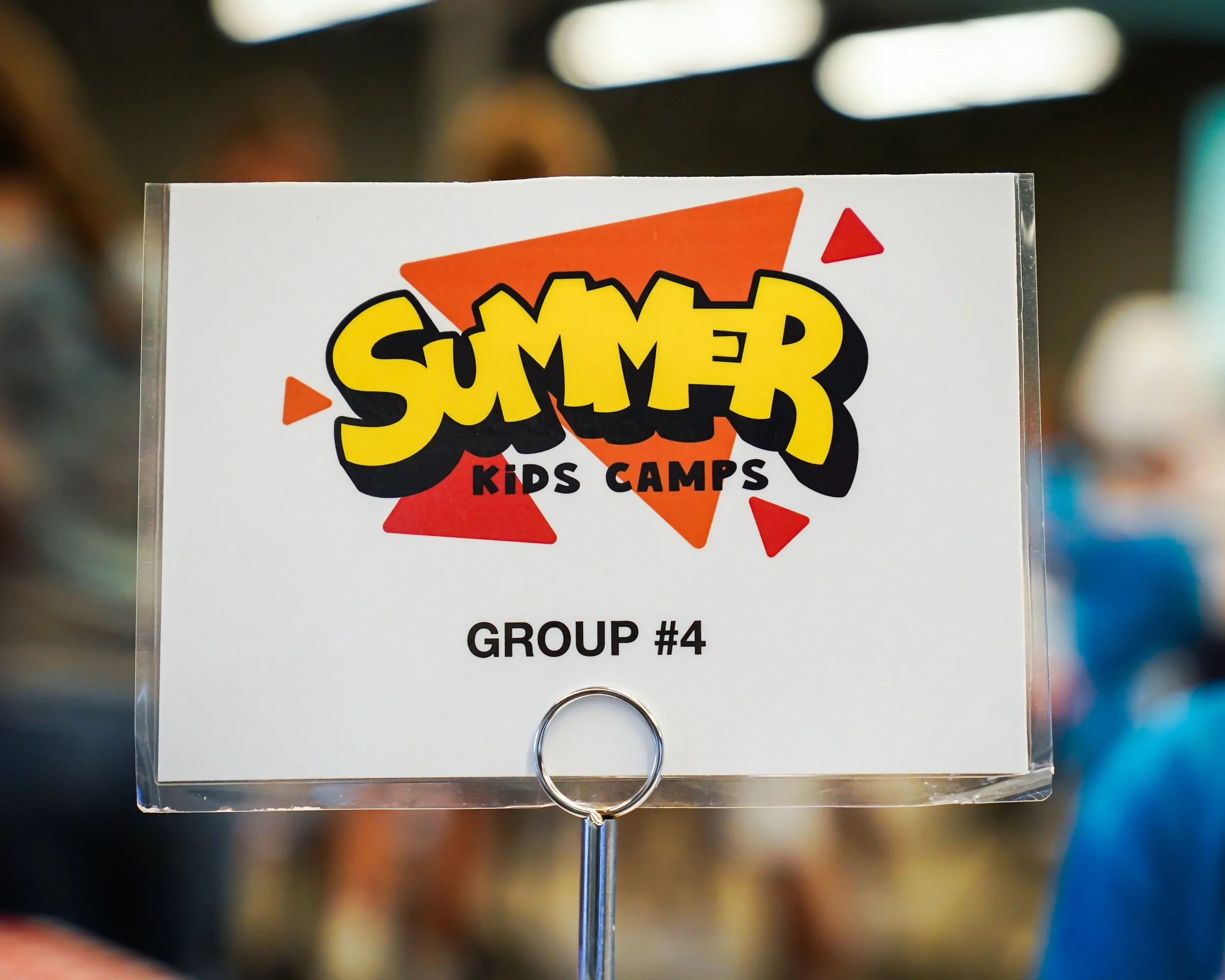 Sign for Summer Kids Camps with group number for an event, featuring colorful graphics and a blurred background.