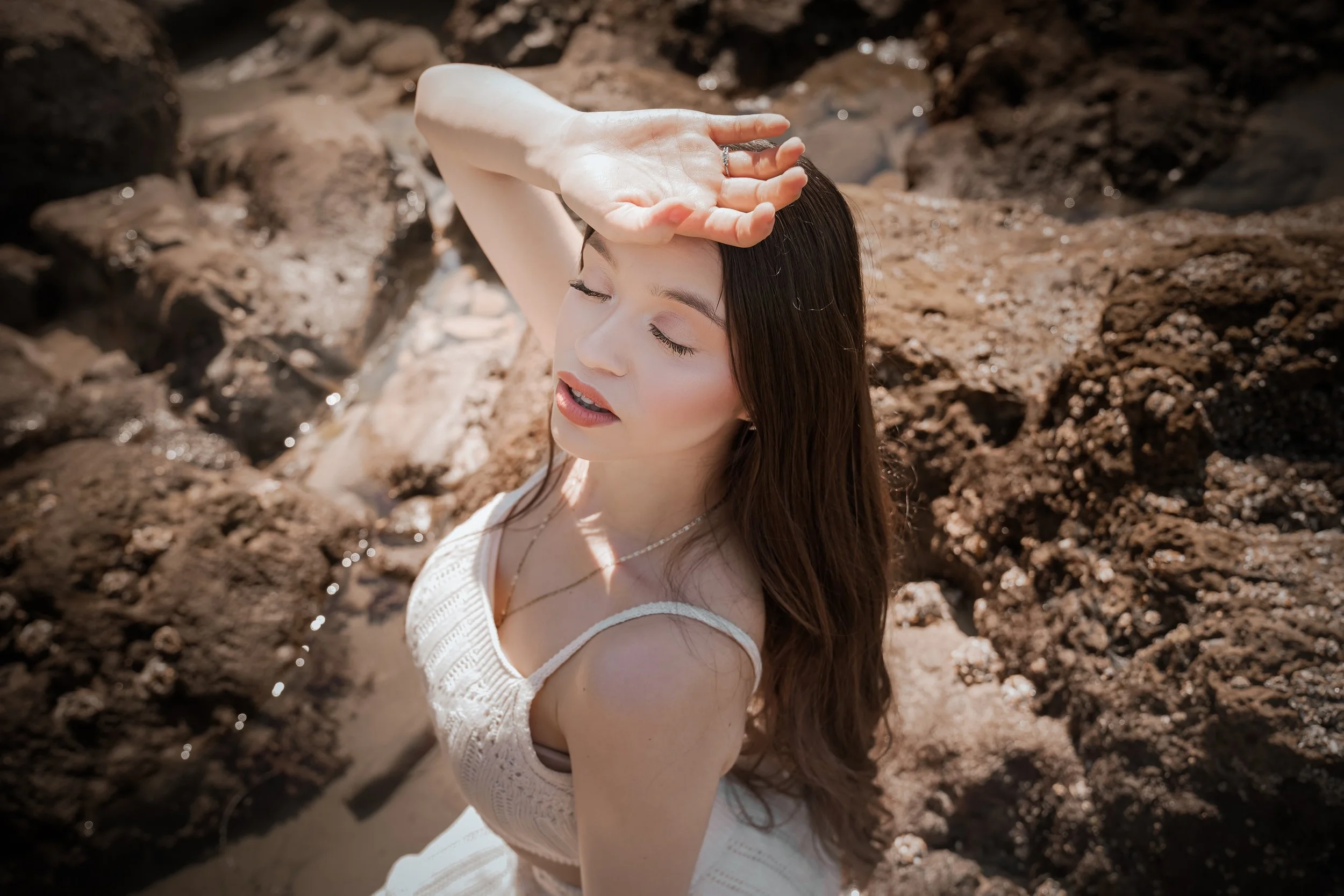 A woman with long brown hair and light skin, dressed in white, sitting on rocks at a beach, with her eyes closed and hand resting on her forehead in bright sunlight.