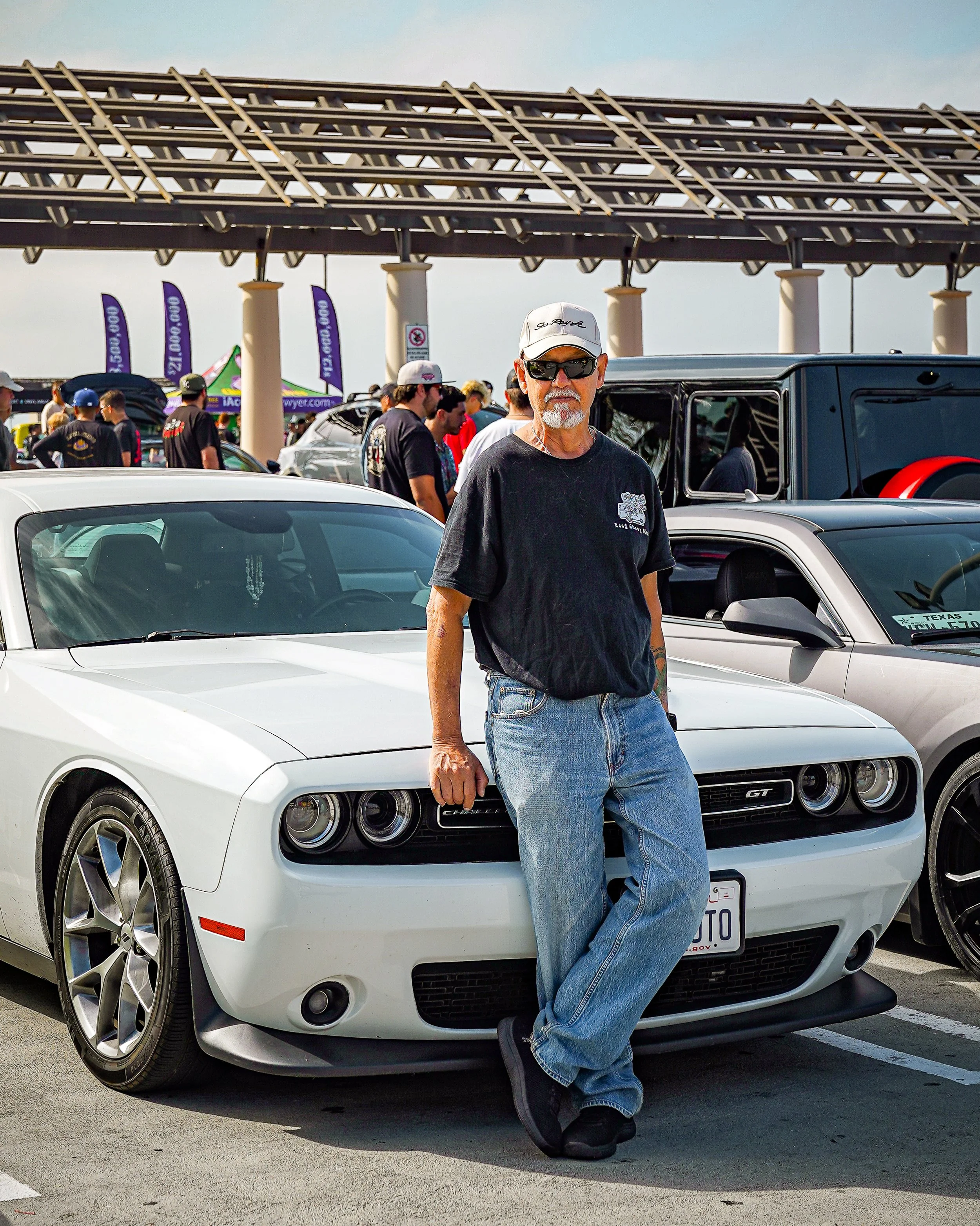 A man with sunglasses, white beard, and gray hair stands next to a white sports car in a crowded car show parking lot.