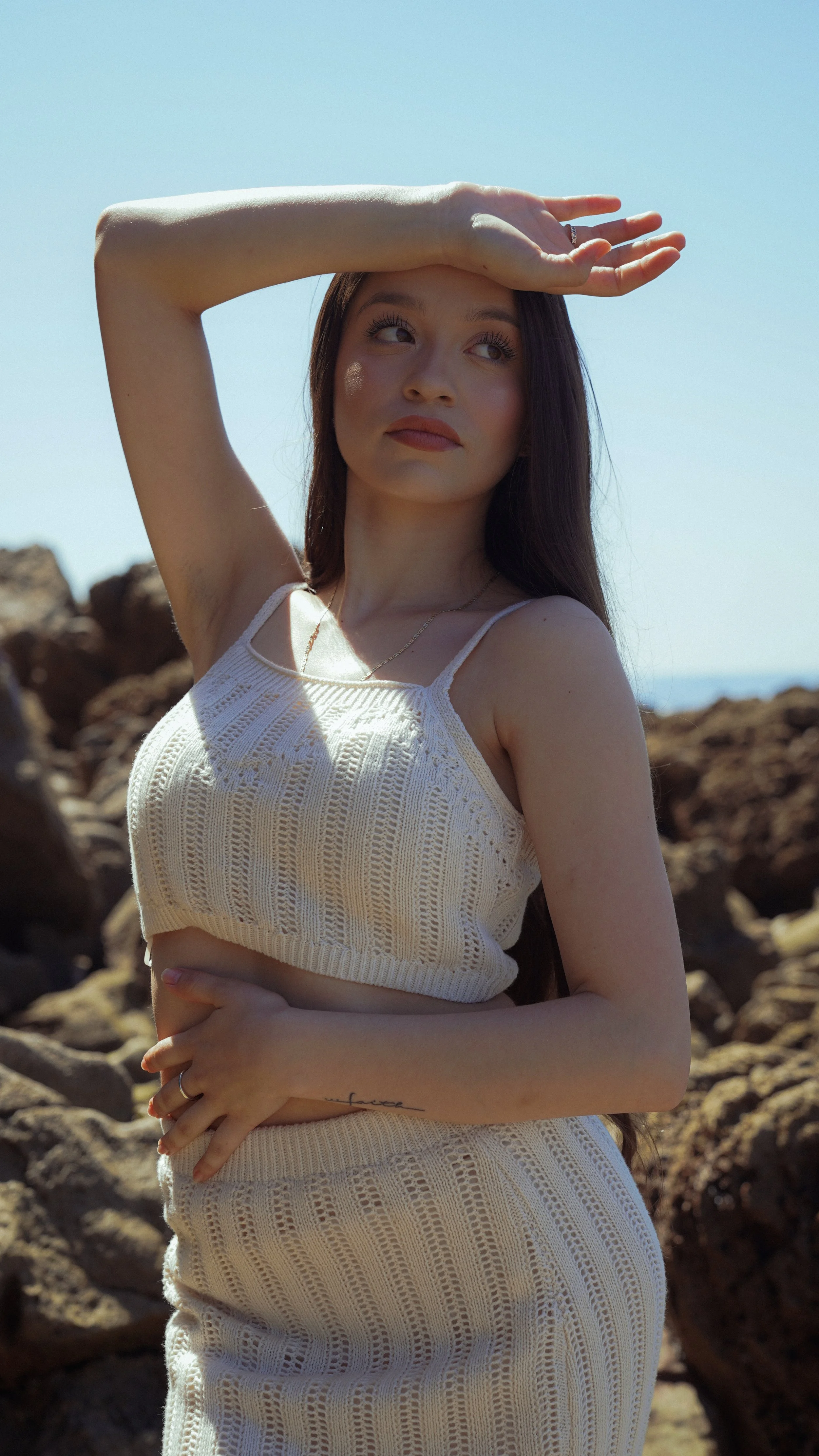 A young woman with long brown hair standing outdoors by rocks, shielding her eyes from the sun with her right hand, wearing a cream-colored knitted tank top and skirt, against a clear blue sky.