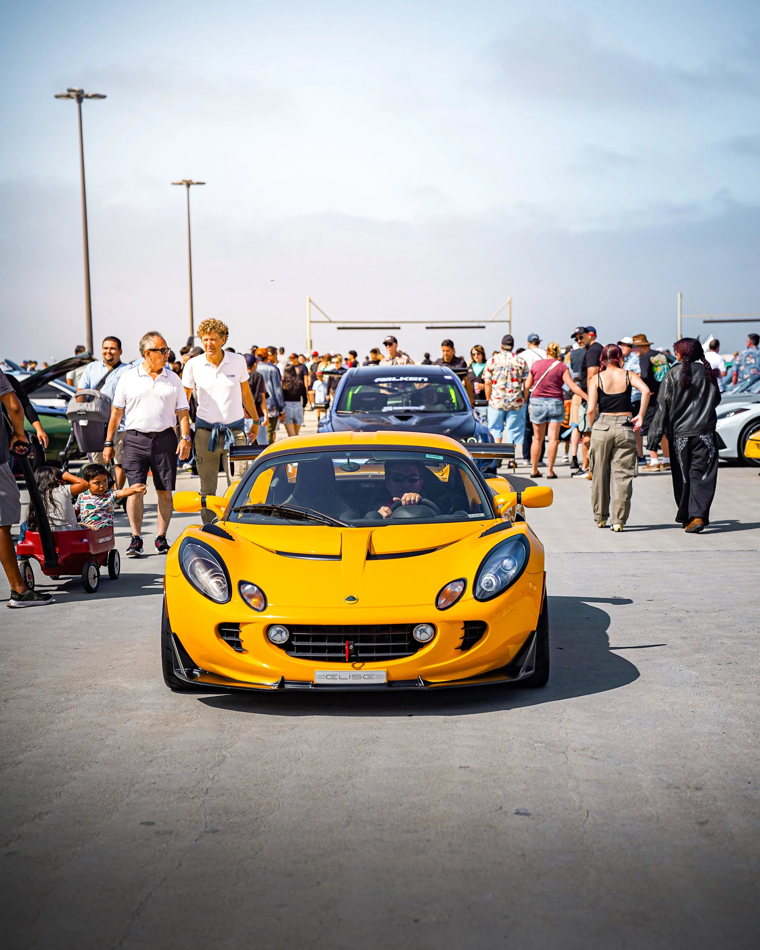 Yellow Lotus Elise sports car at a car show with a crowd of people walking around.